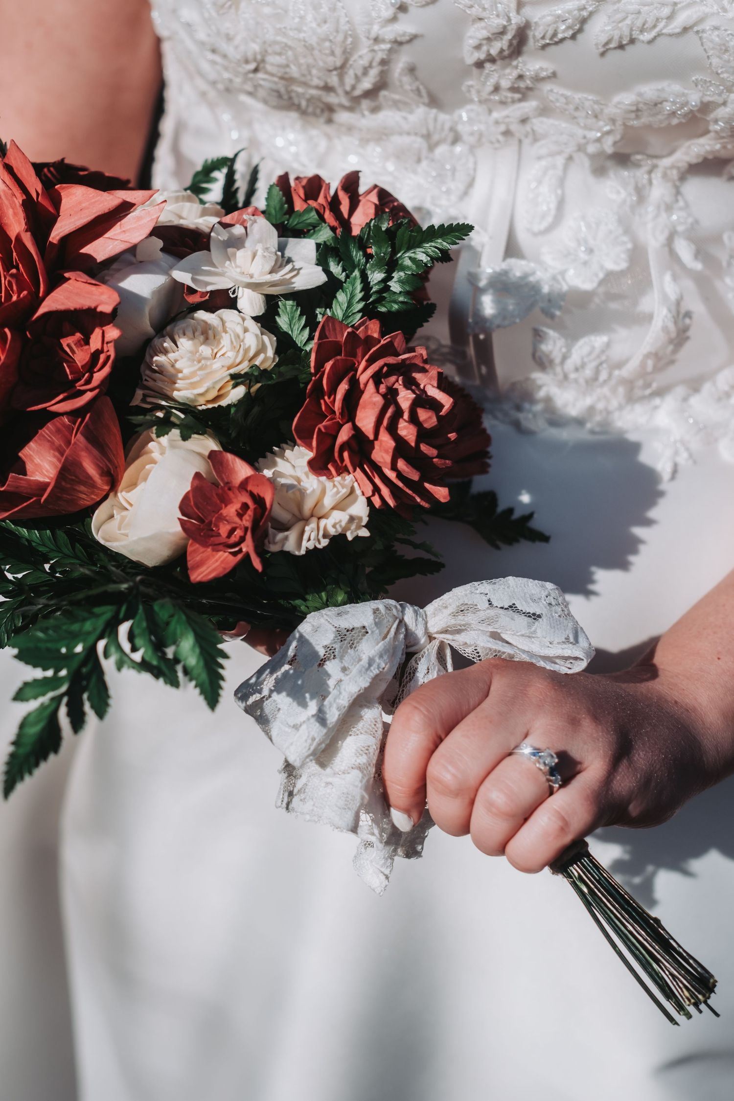 Bride holding a wedding bouquet of red and white flowers with lace ribbon, wearing an engagement ring and white lace gown.