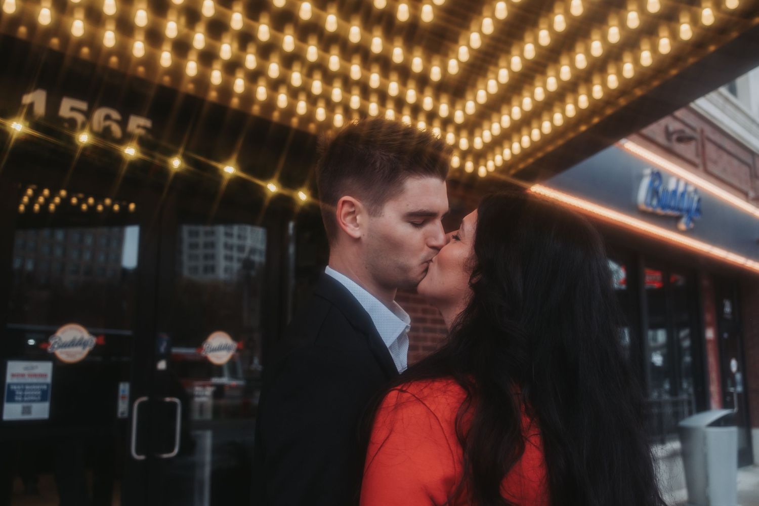 Couple sharing a romantic kiss under glowing marquee lights outside a city venue at night, woman in red dress.