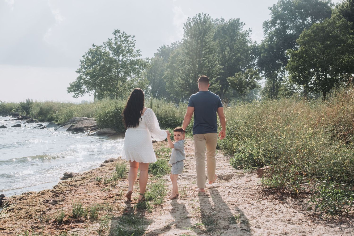 Family of three walking hand-in-hand along a sunny lakeside path surrounded by lush greenery.