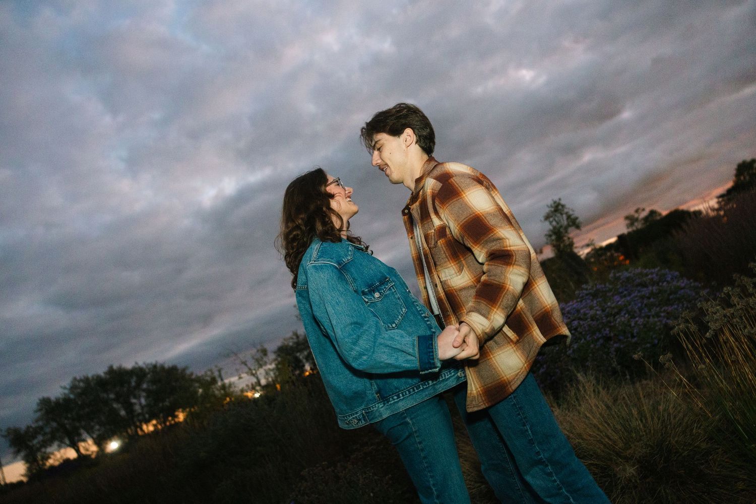 Couple holding hands at dusk under dramatic cloudy sky, wearing casual denim and plaid outfits in an outdoor setting.