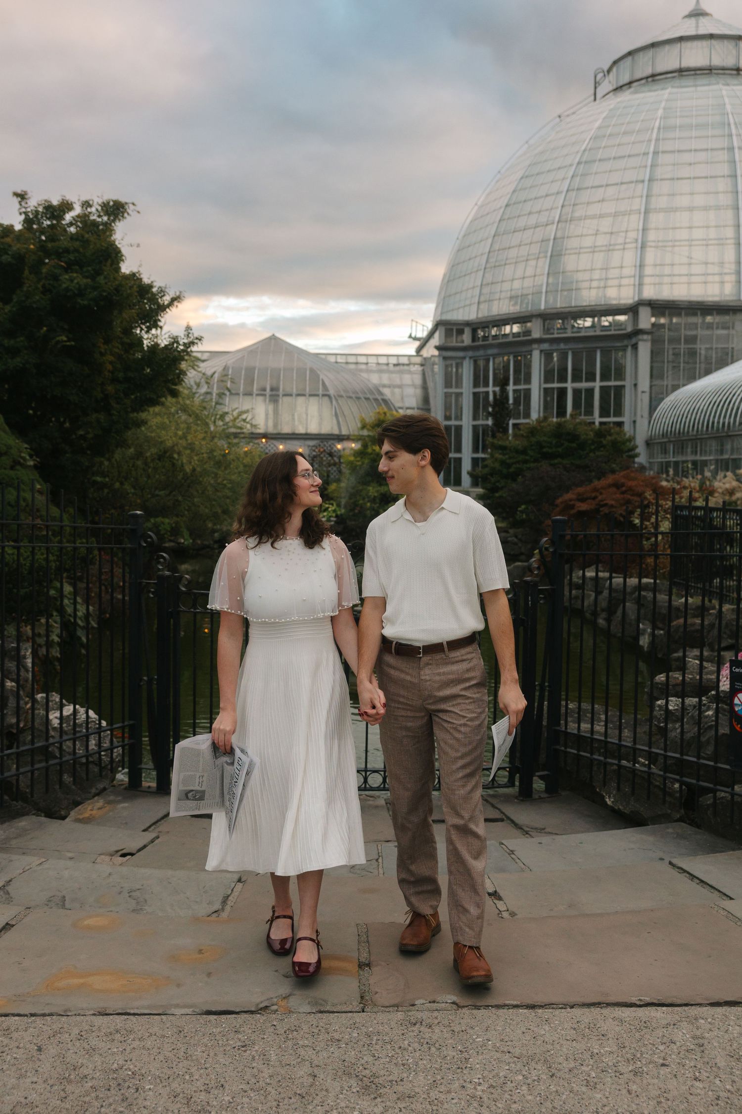 A couple holds hands and gazes at each other in front of a glass greenhouse dome at a botanical garden.