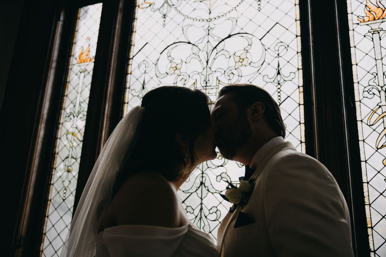 Silhouette of bride and groom sharing a kiss in front of ornate stained glass windows on their wedding day.