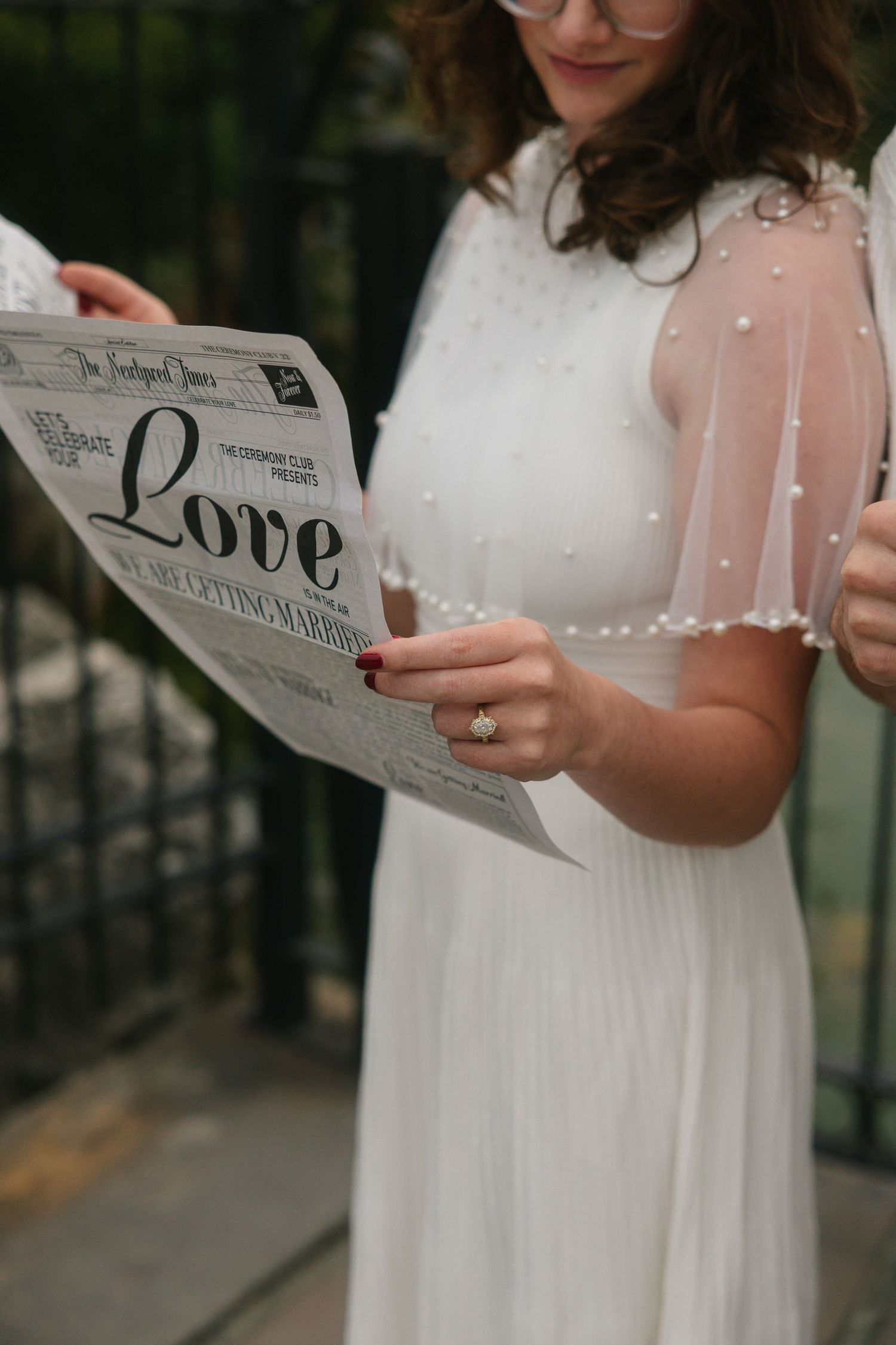Bride in white pearl-embellished dress reading a Love newspaper, showcasing her diamond engagement ring.