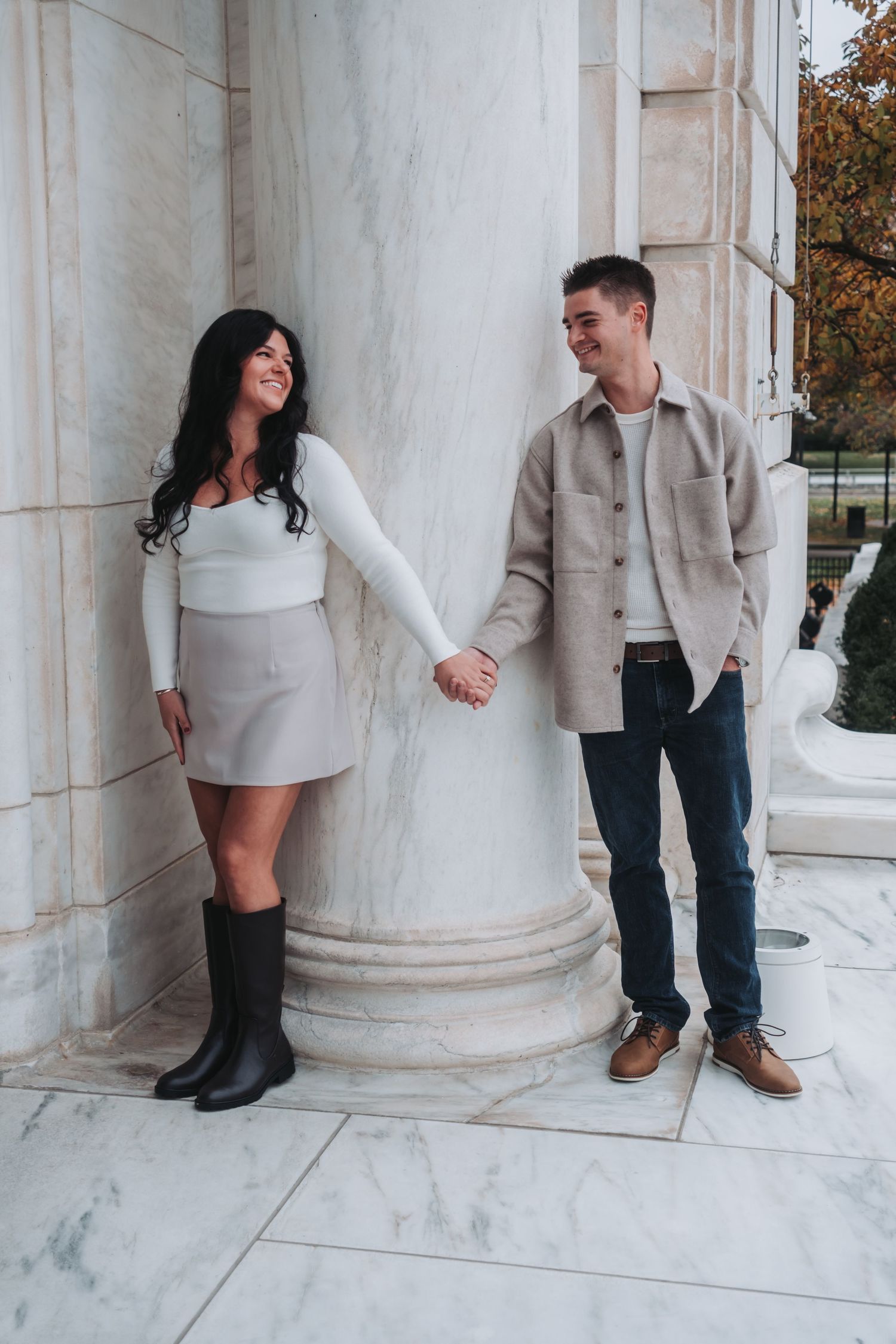 A couple holds hands and smiles at each other beside a marble column, dressed in casual chic outfits.