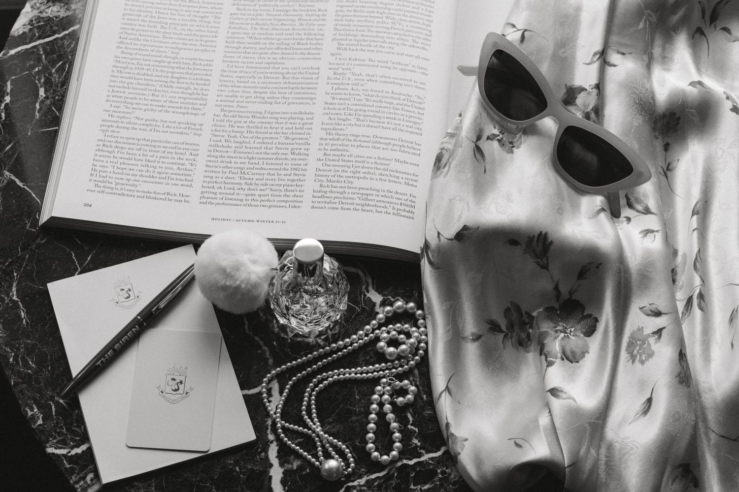 Flatlay of an open book, sunglasses, jewelry and decorative items on dark marble surface.