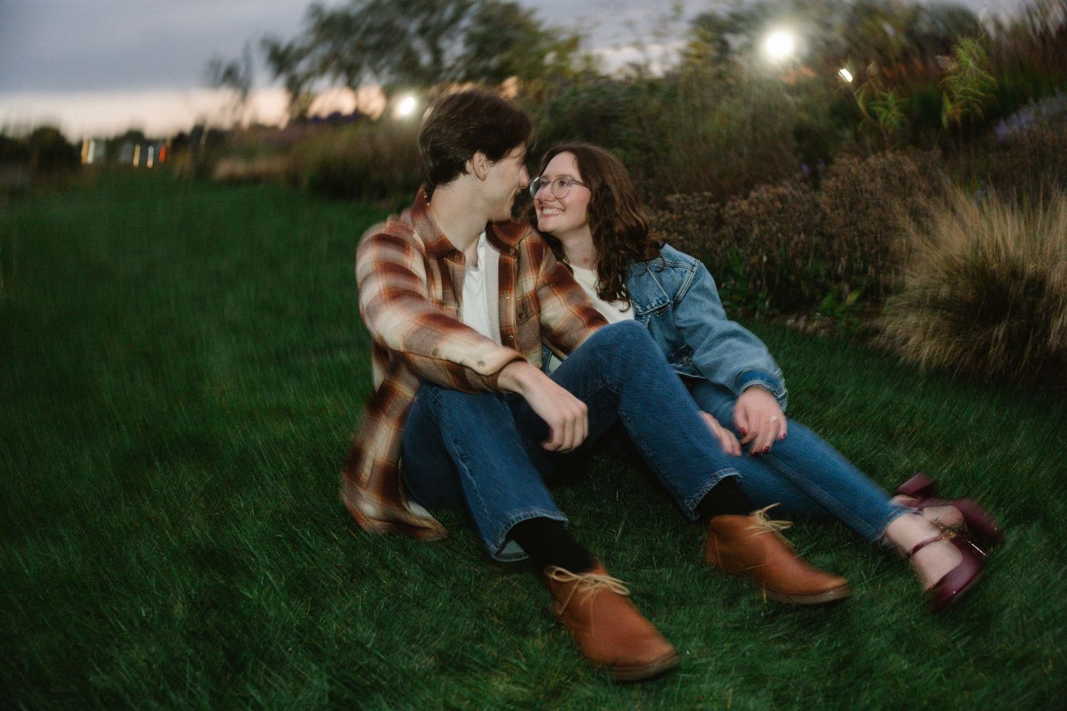 Couple sitting on green grass at dusk, wearing flannel shirts and jeans, smiling at each other romantically.
