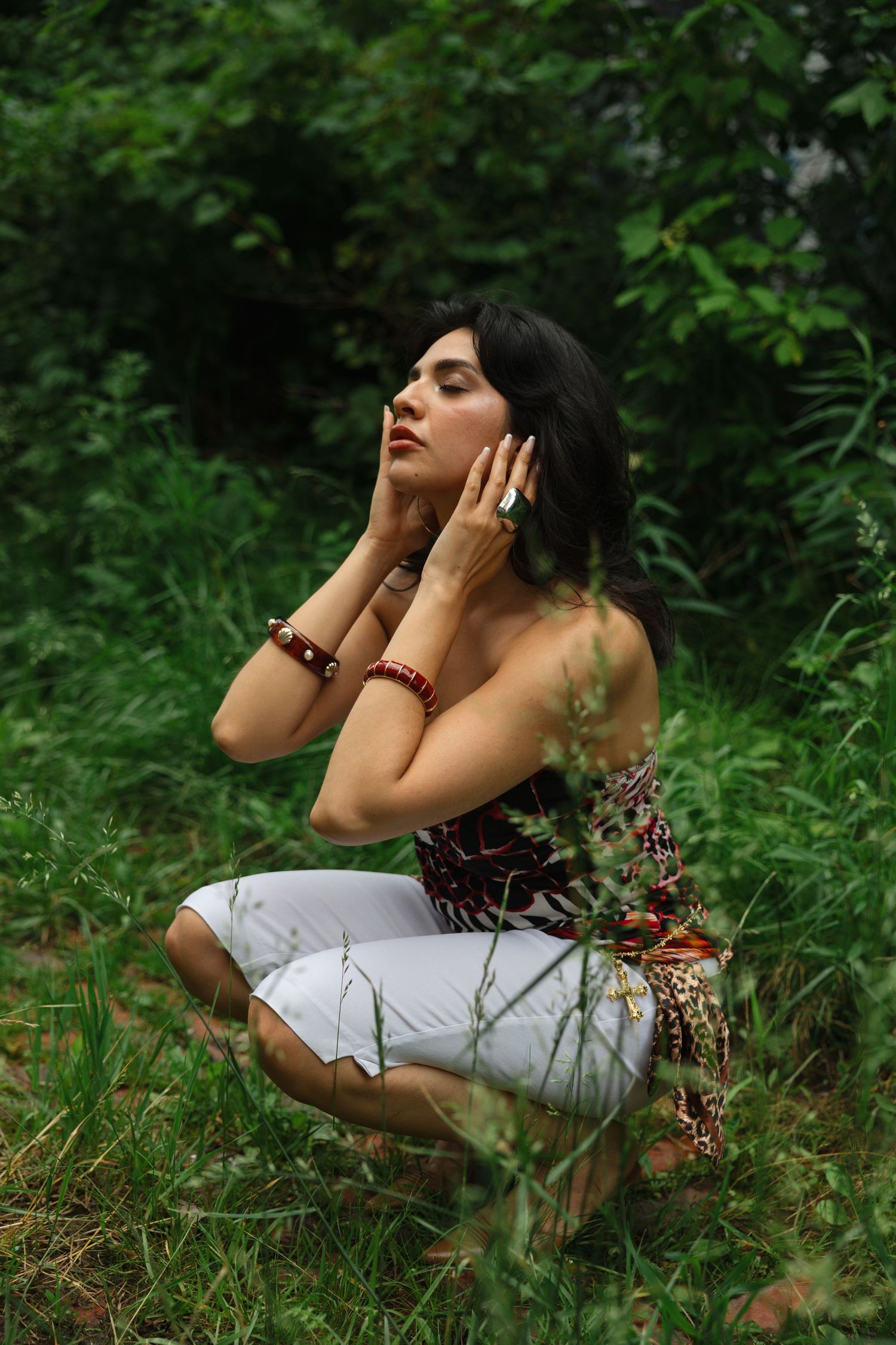 Woman in floral top and white pants crouching in lush green forest, hands framing face, wearing red bracelets.