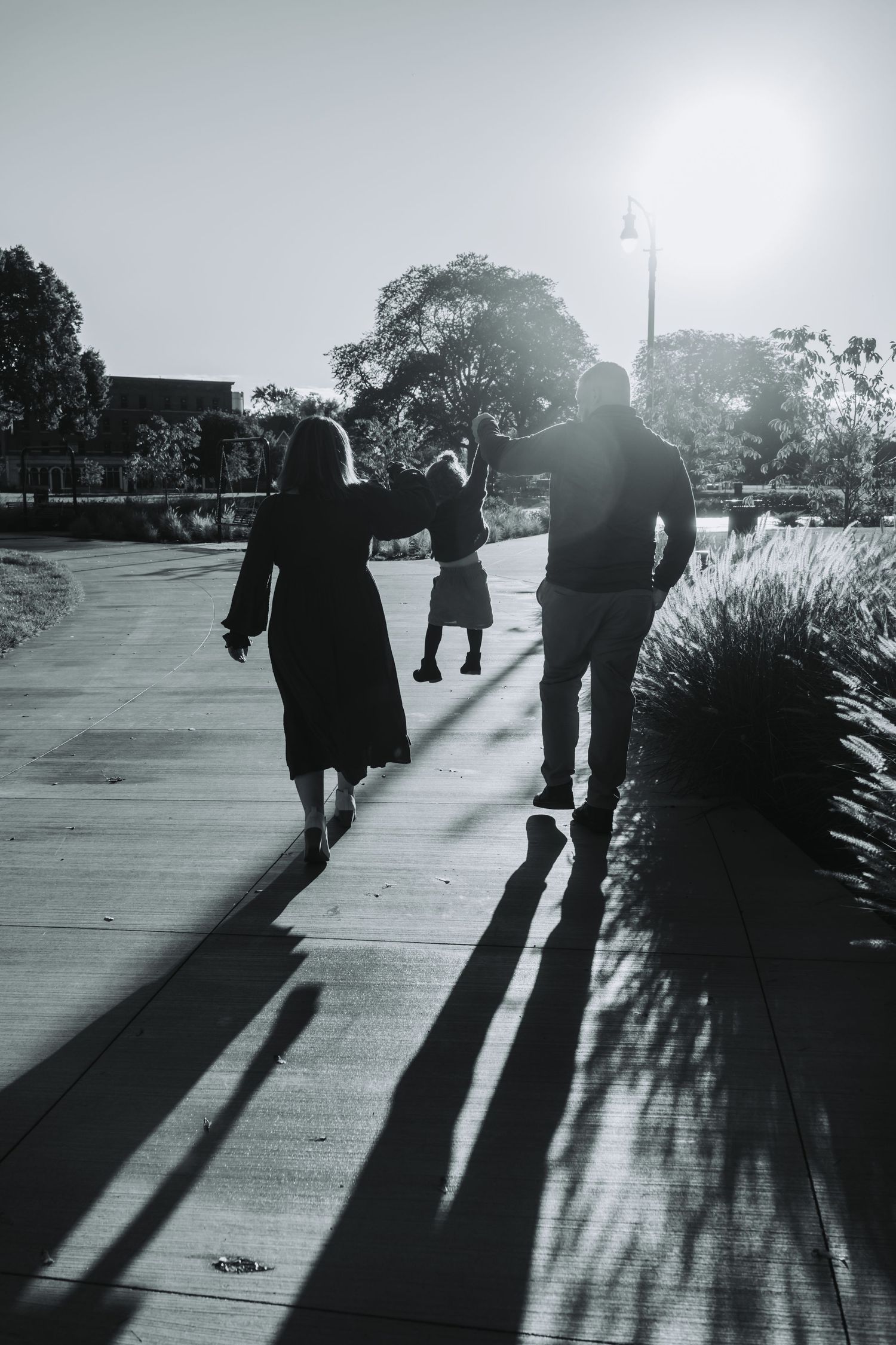 Black and white photo of two people walking on a sunlit path, casting long shadows, with trees and a lamppost in the background.