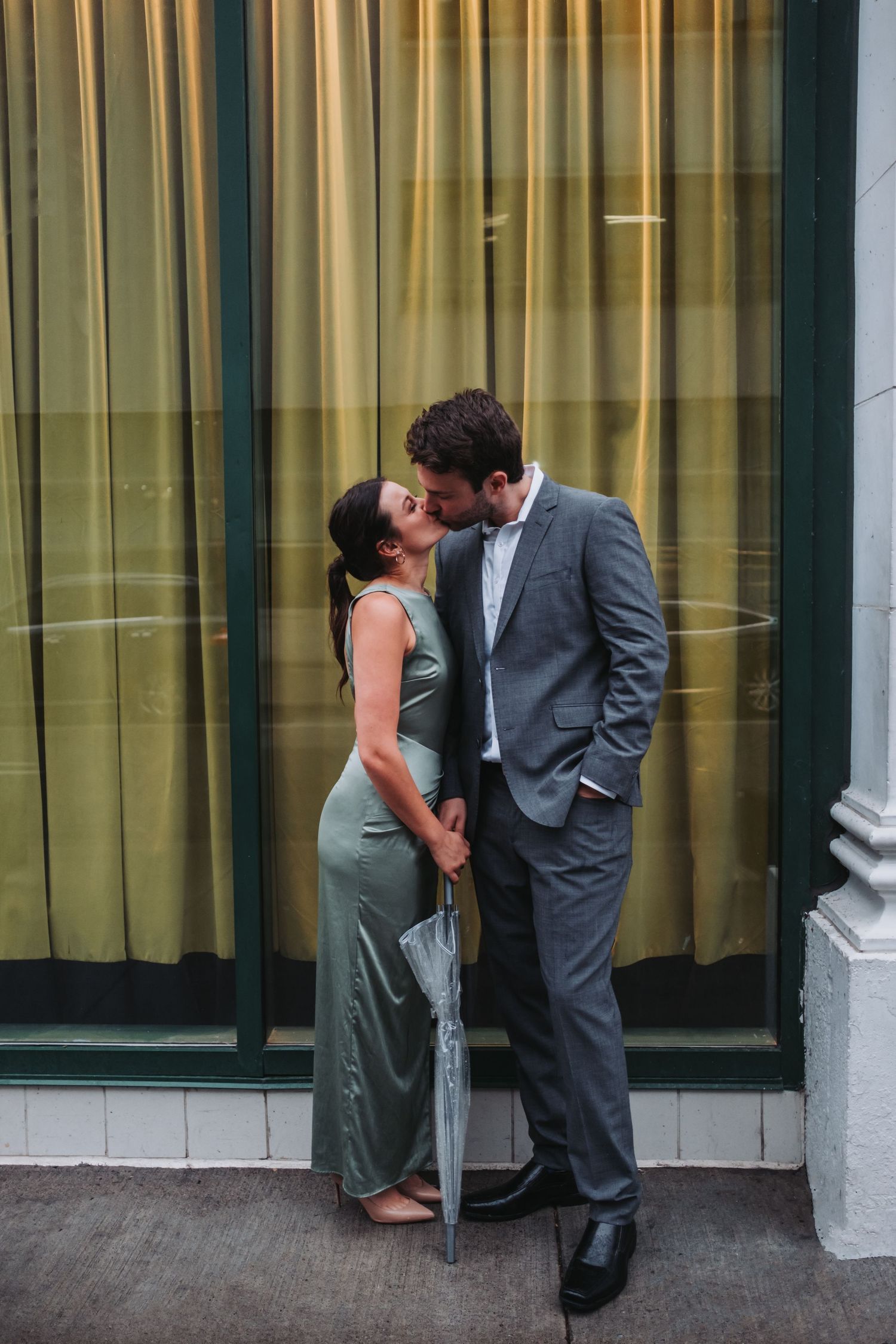 Couple kissing in elegant attire, woman in green dress, man in gray suit, holding umbrella by yellow curtains.