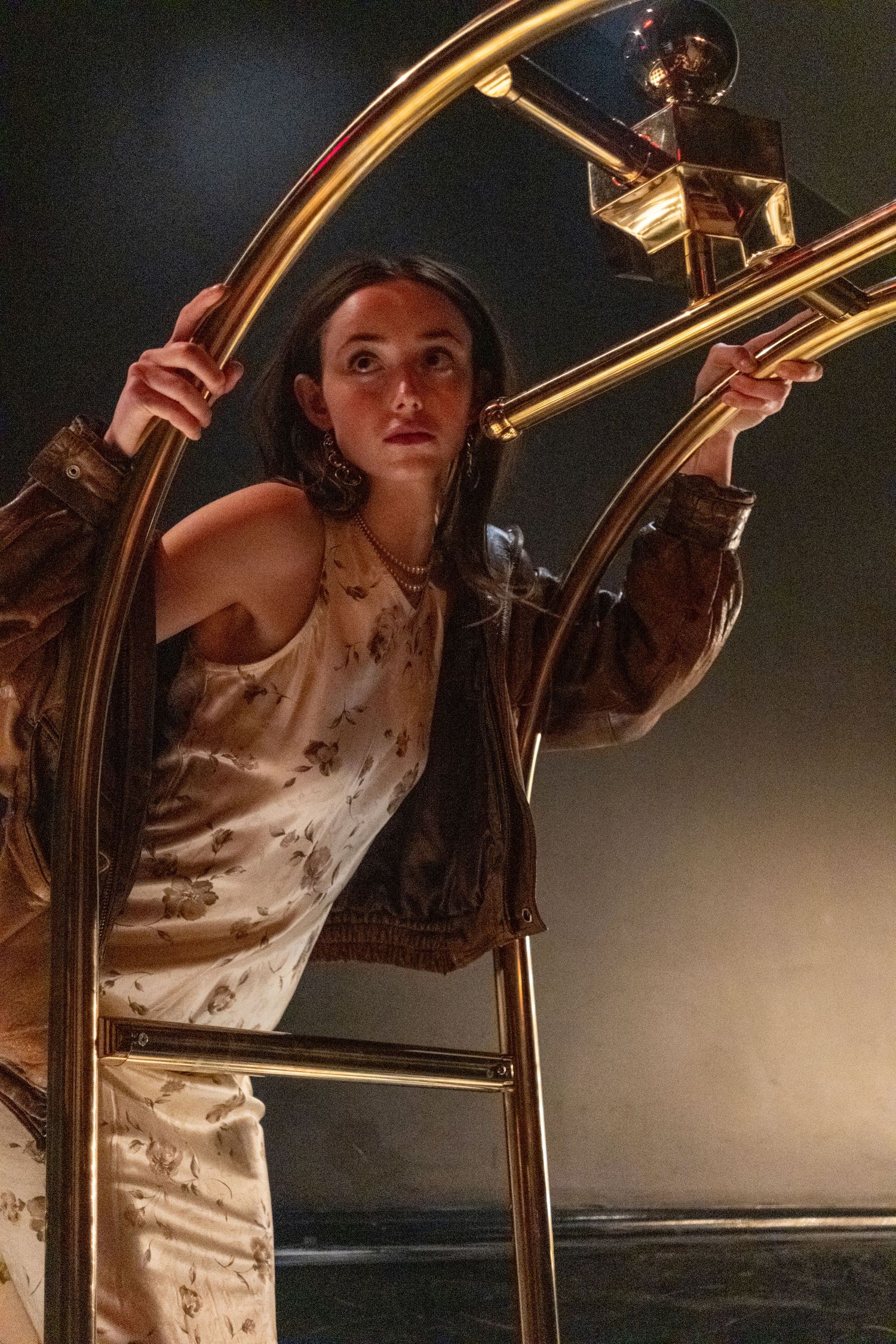 Young woman in satin dress and brown jacket poses dramatically with gold luggage cart in moody lighting.