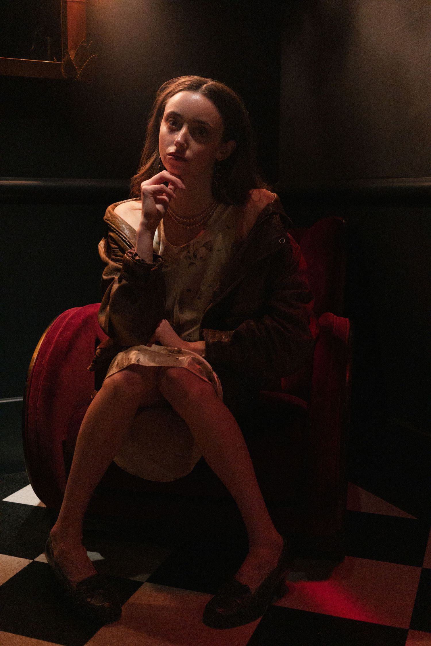 A woman in a floral dress and brown jacket sits pensively in a red velvet chair on a checkered floor in dim lighting.