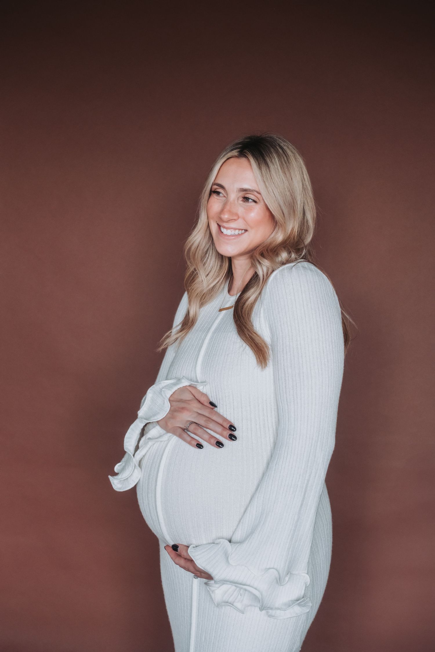 Smiling pregnant woman in white ribbed dress cradling baby bump against brown background.