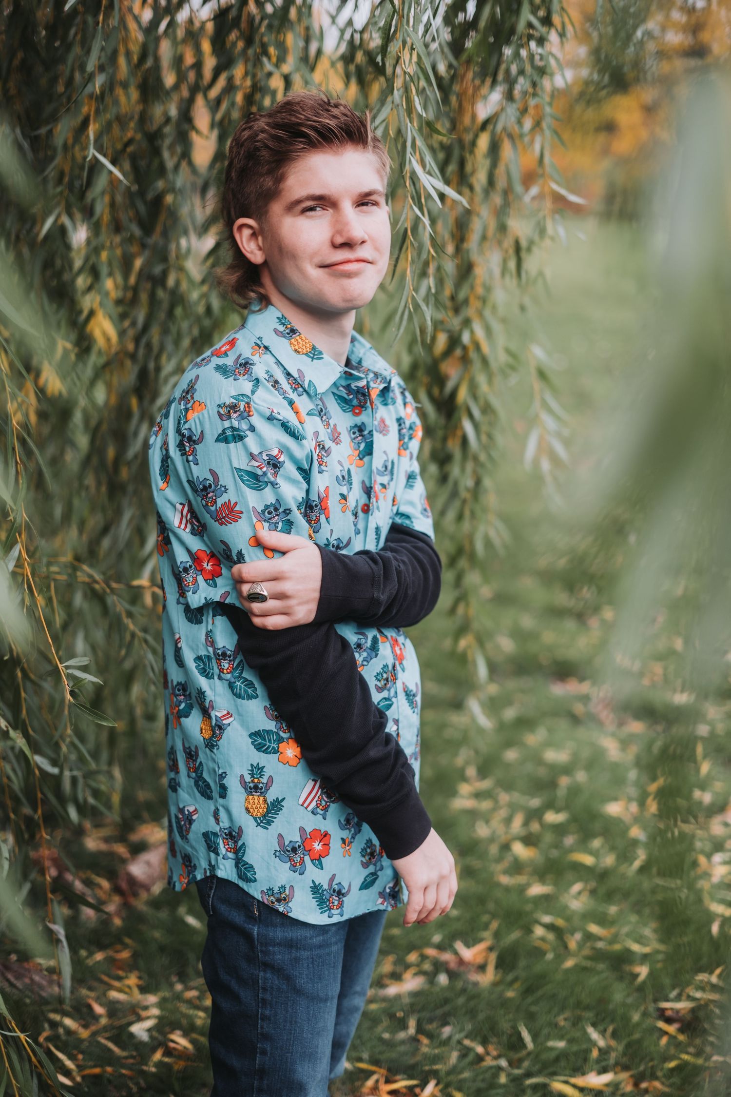 Young person in floral button-up shirt and black sleeves poses against willow tree branches.