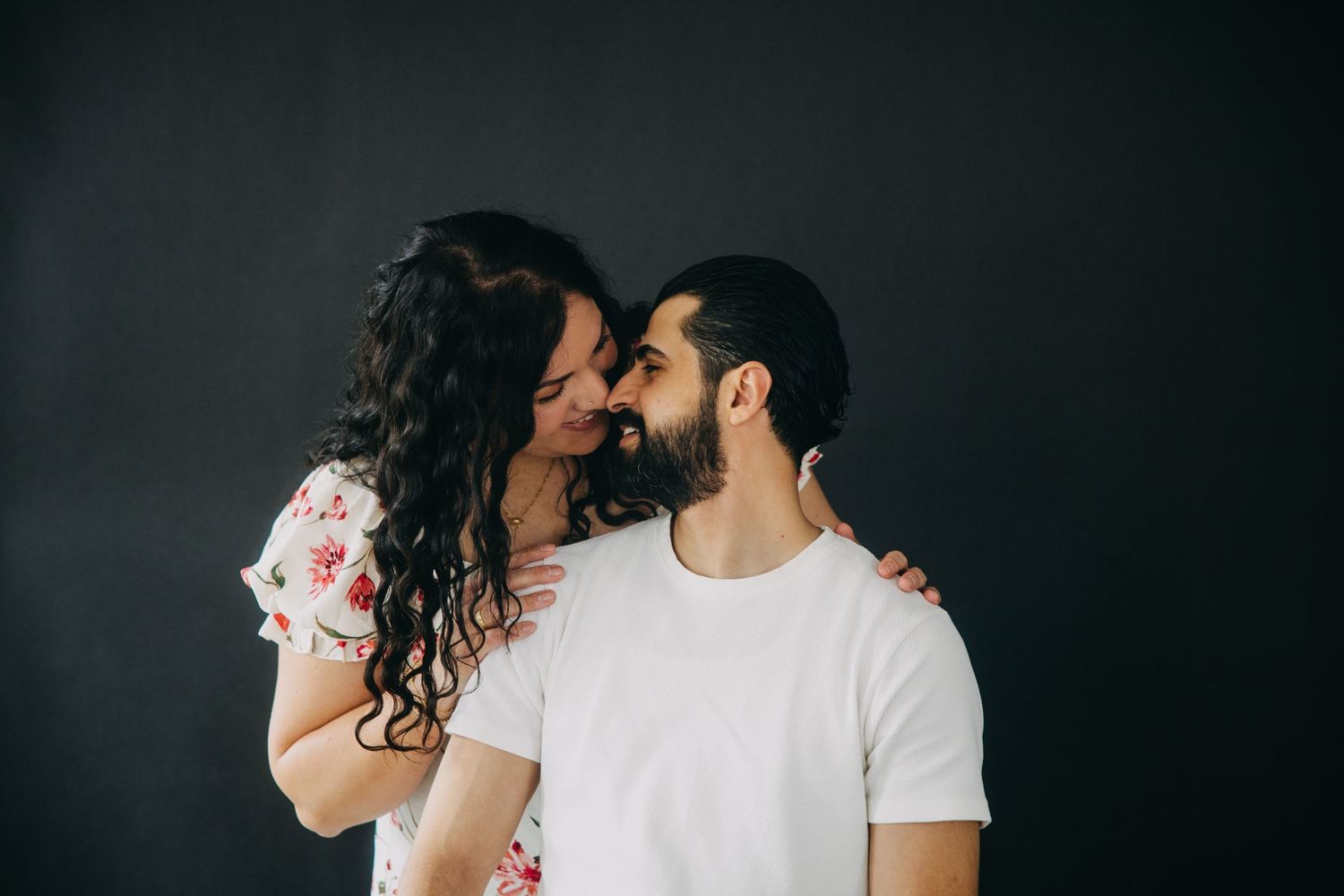 Romantic couple sharing an intimate moment, woman in floral dress embracing bearded man in white tee against dark background.