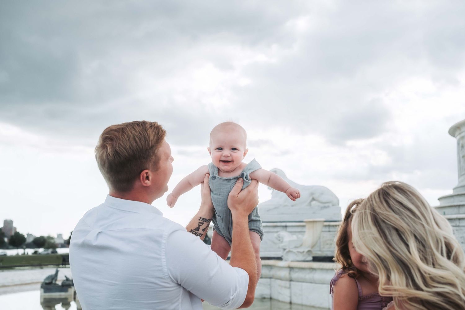 Parents joyfully lifting smiling baby outdoors against cloudy sky near waterfront.