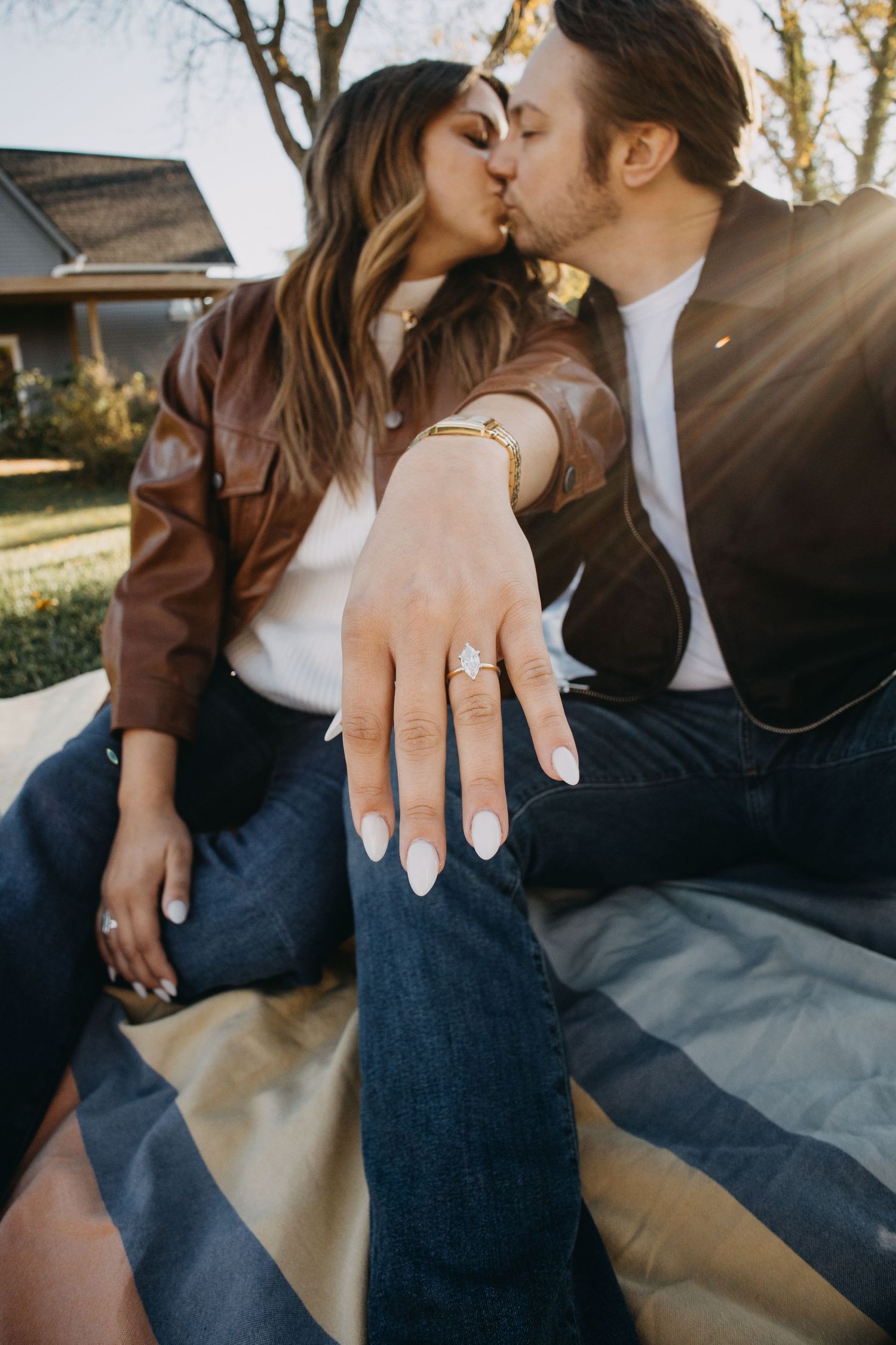 Couple kissing outdoors while showing off engagement ring with diamond solitaire, both wearing leather jackets.