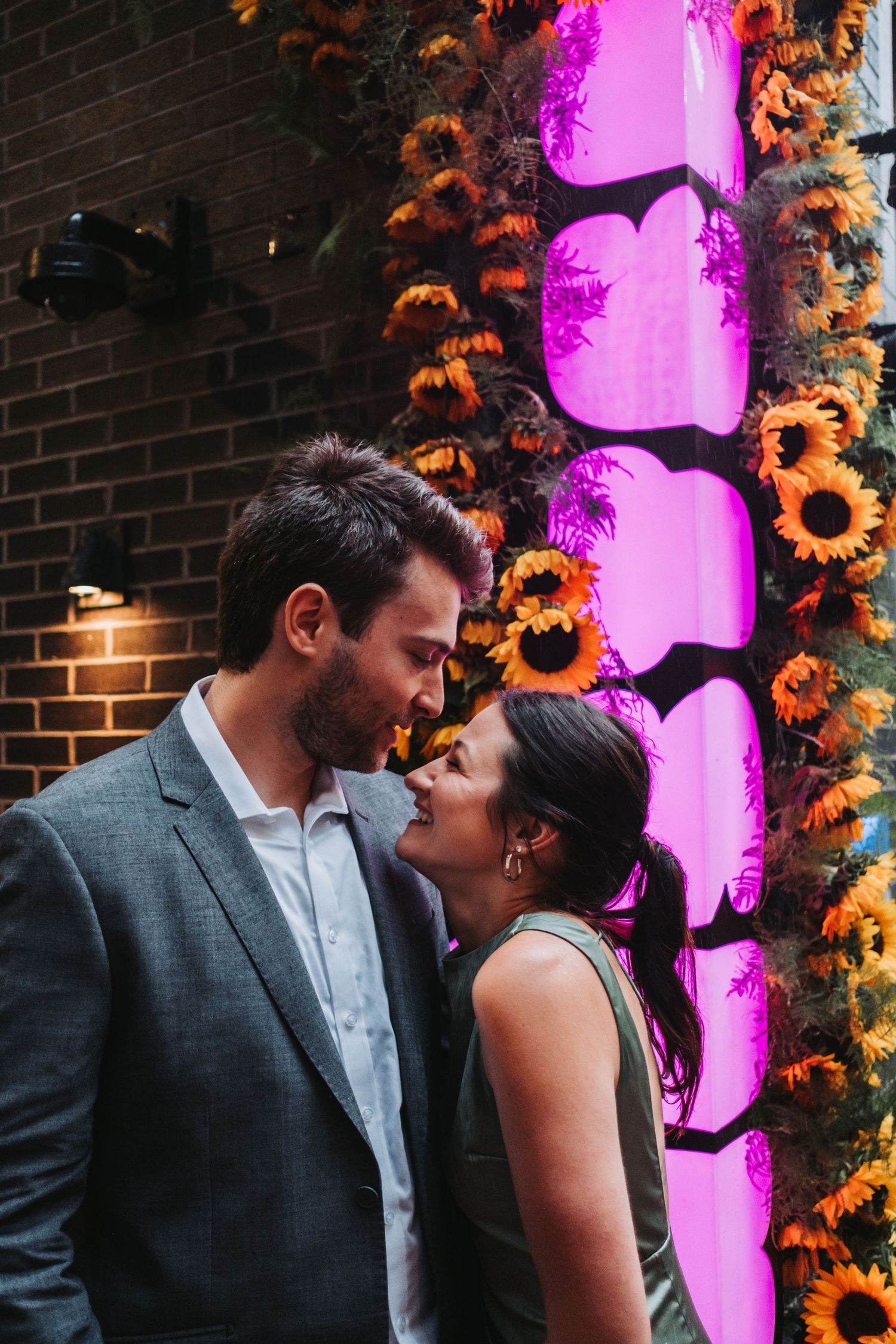 Couple sharing a romantic moment in front of a vibrant pink floral installation adorned with sunflowers.