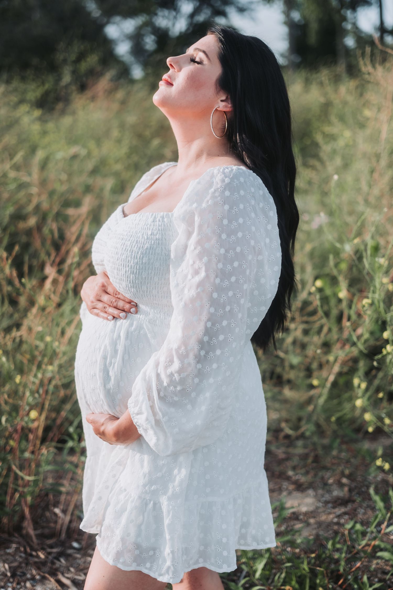 Pregnant woman in white polka dot dress cradling baby bump outdoors in natural field setting.