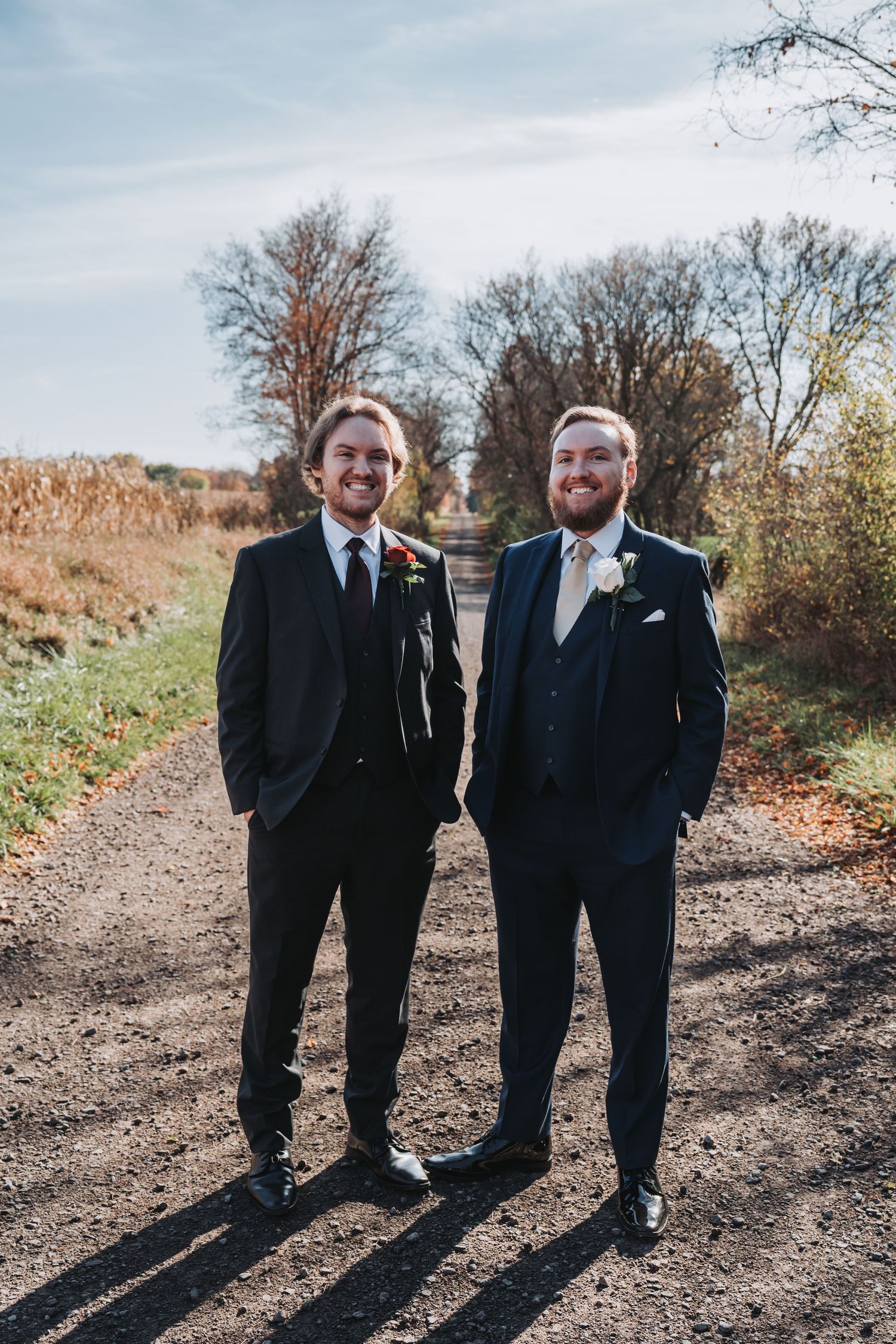 Two grooms in dark suits with boutonnieres stand smiling on a gravel path surrounded by autumn trees.