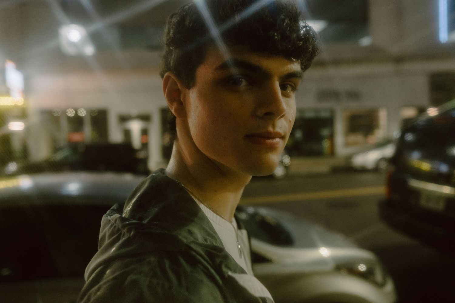 Young person in a green jacket standing outdoors at night with cars and city lights in the background.