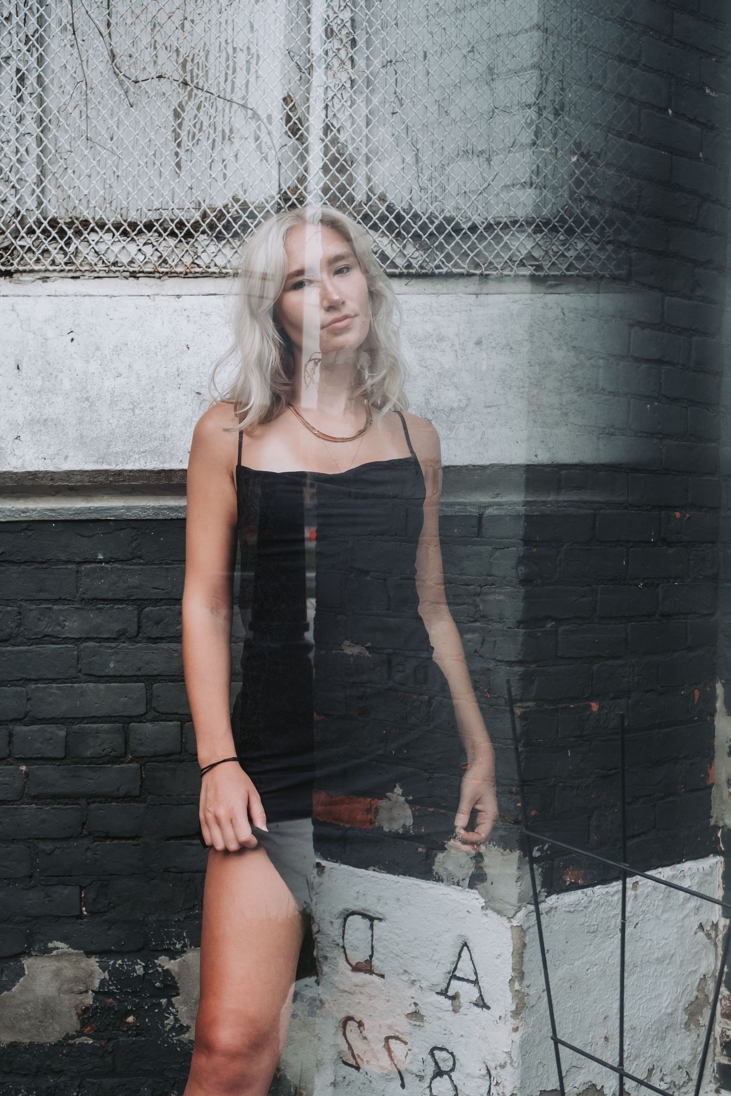 Young woman with blonde hair in black mini dress posing against graffiti brick wall in urban setting.