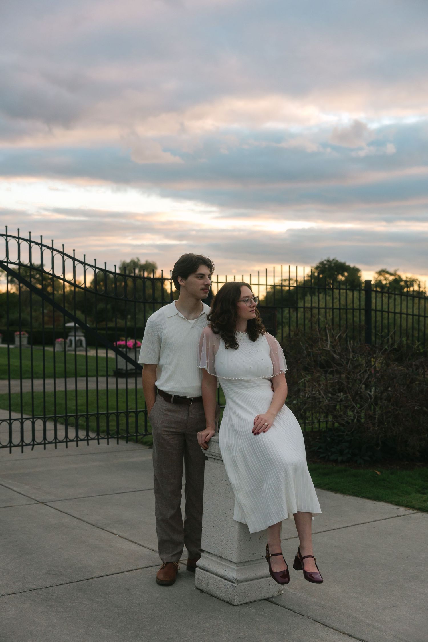A couple poses at sunset near iron gates, woman in white dress, man in khakis, with dramatic cloudy sky backdrop.