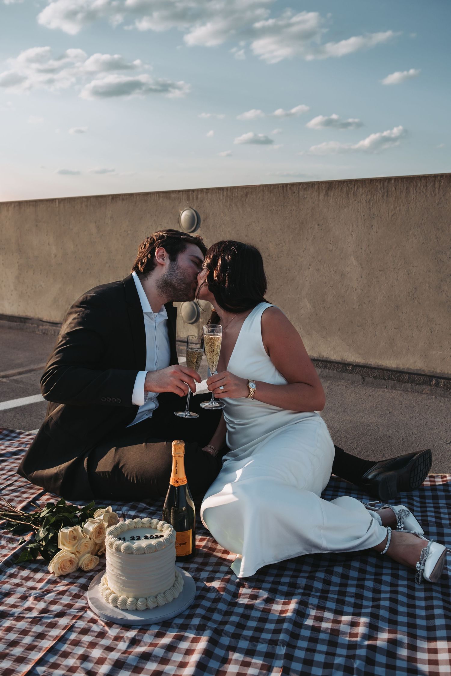 Couple kissing on rooftop picnic blanket with champagne, wedding cake, and roses at sunset celebration.