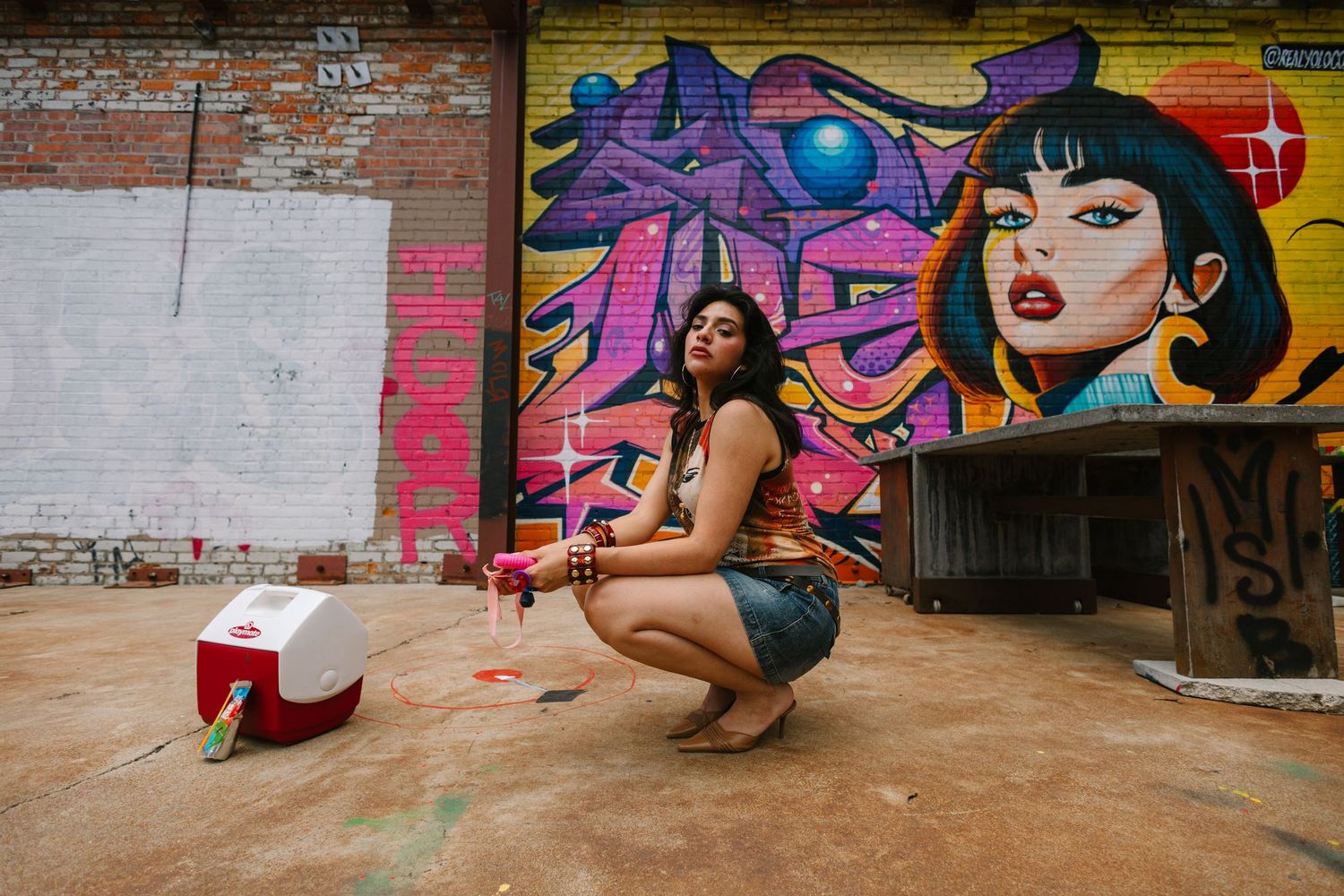 Woman crouching near cooler in front of vibrant graffiti mural outdoors.