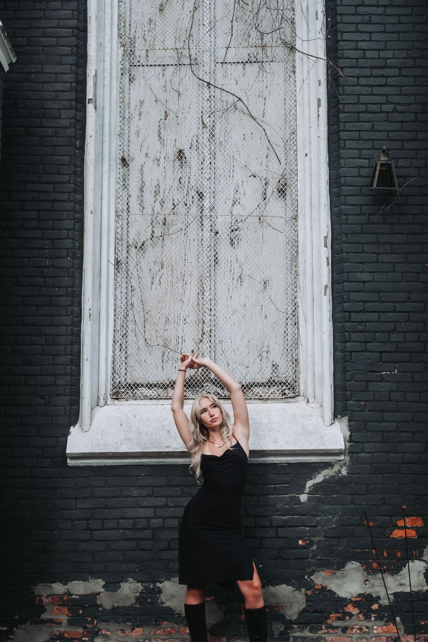 A figure in black poses against a weathered white door on dark brick wall.