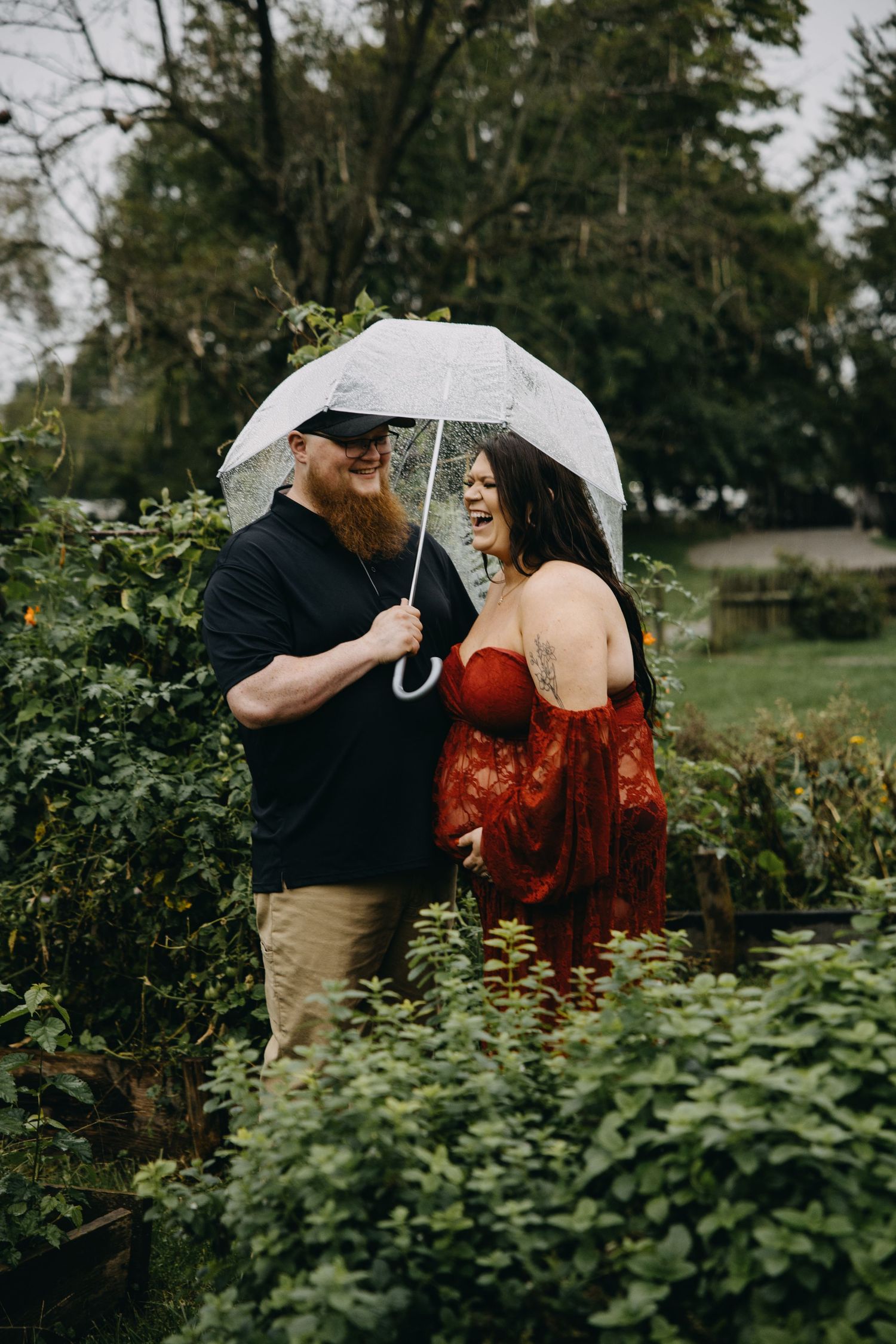 Couple laughing under clear umbrella in lush garden, man with red beard in black shirt, woman in red maternity gown.
