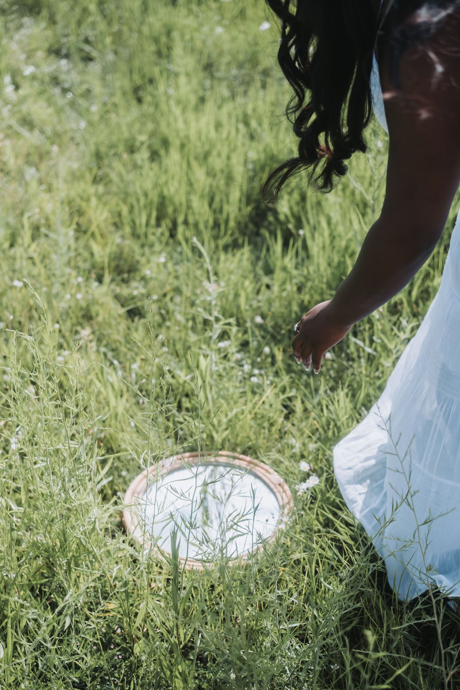 Woman in white dress bending over a round mirror placed in lush green grass outdoors.