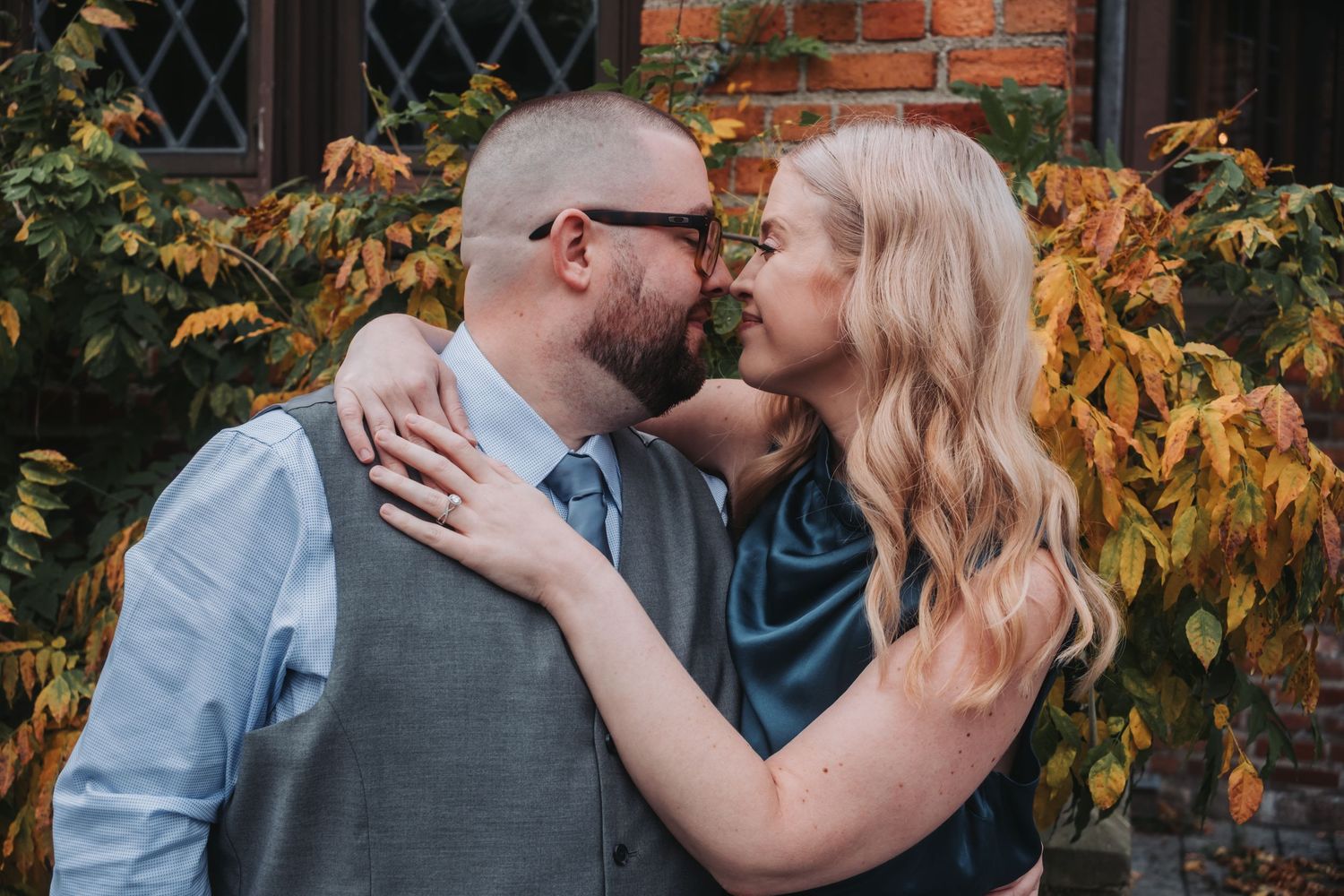Couple sharing a romantic moment outdoors, surrounded by autumn foliage near a brick wall.