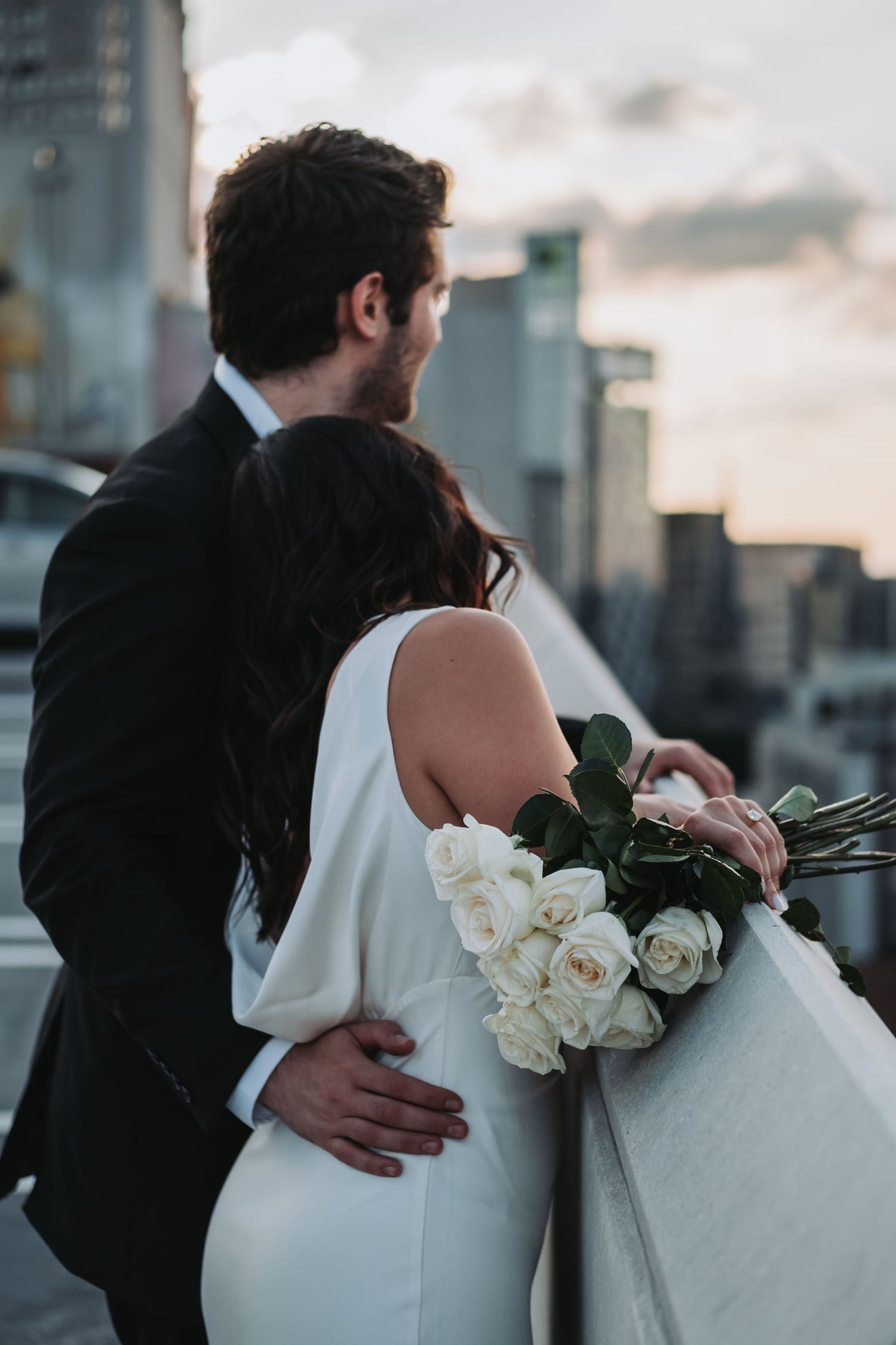 Bride and groom stand on rooftop at sunset, she holds white roses bouquet as he embraces her waist with city skyline backdrop.