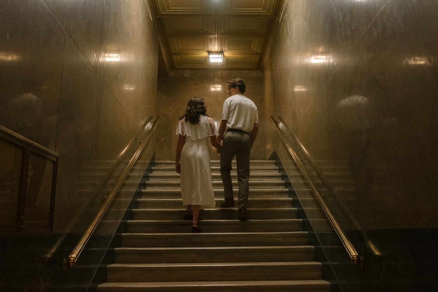 Couple holding hands walking up grand staircase in elegant golden-lit hall, viewed from behind.