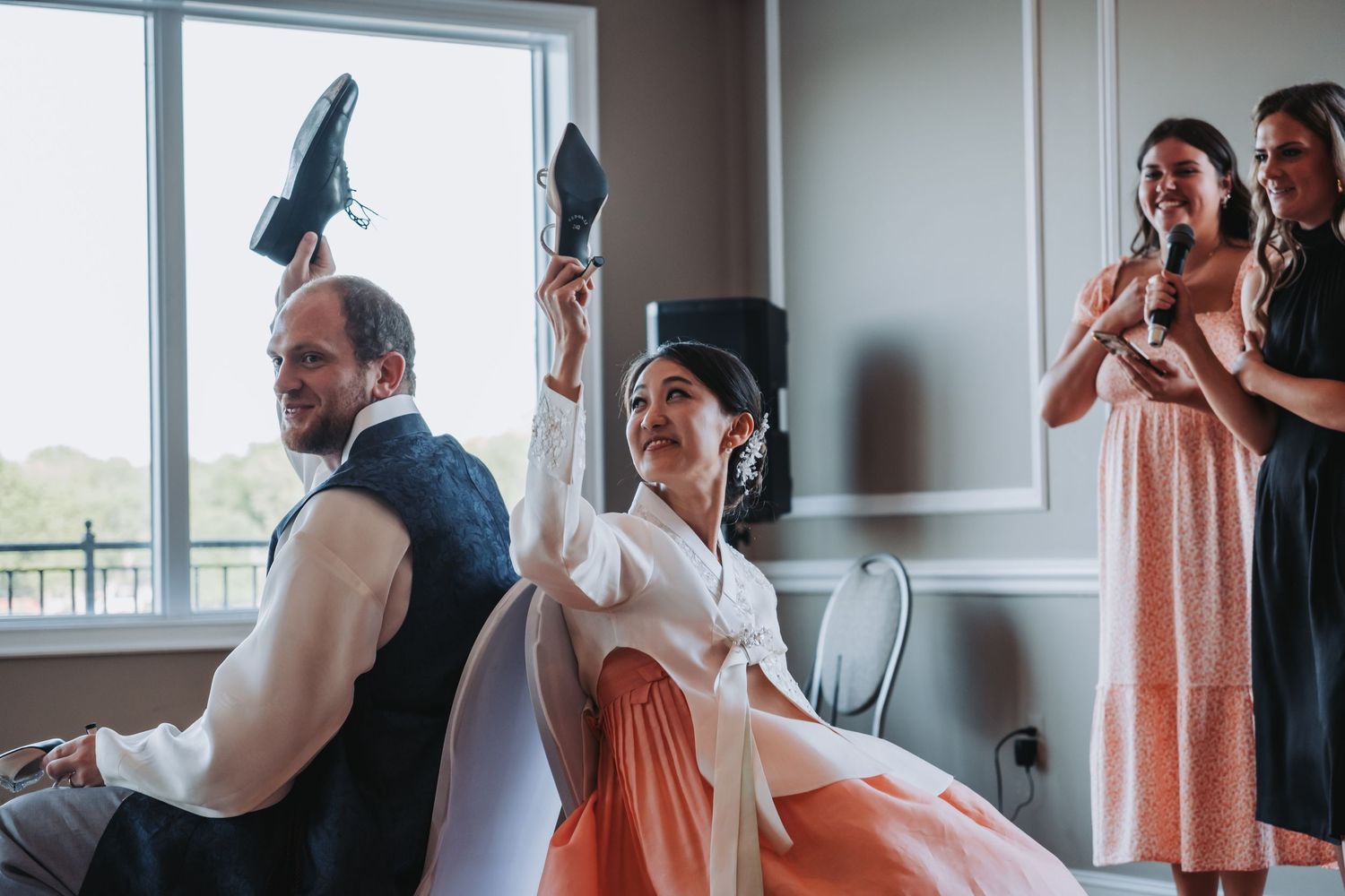 Wedding guests in peach-colored dresses gather by large windows in an indoor venue.