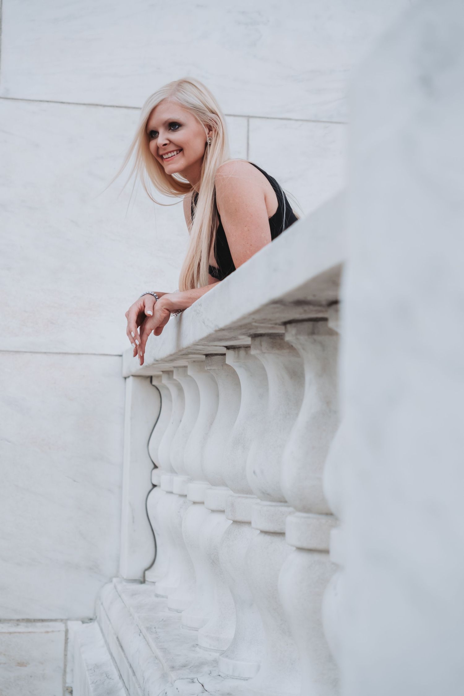 Smiling blonde woman in black dress leaning on white marble balustrade railing outdoors.