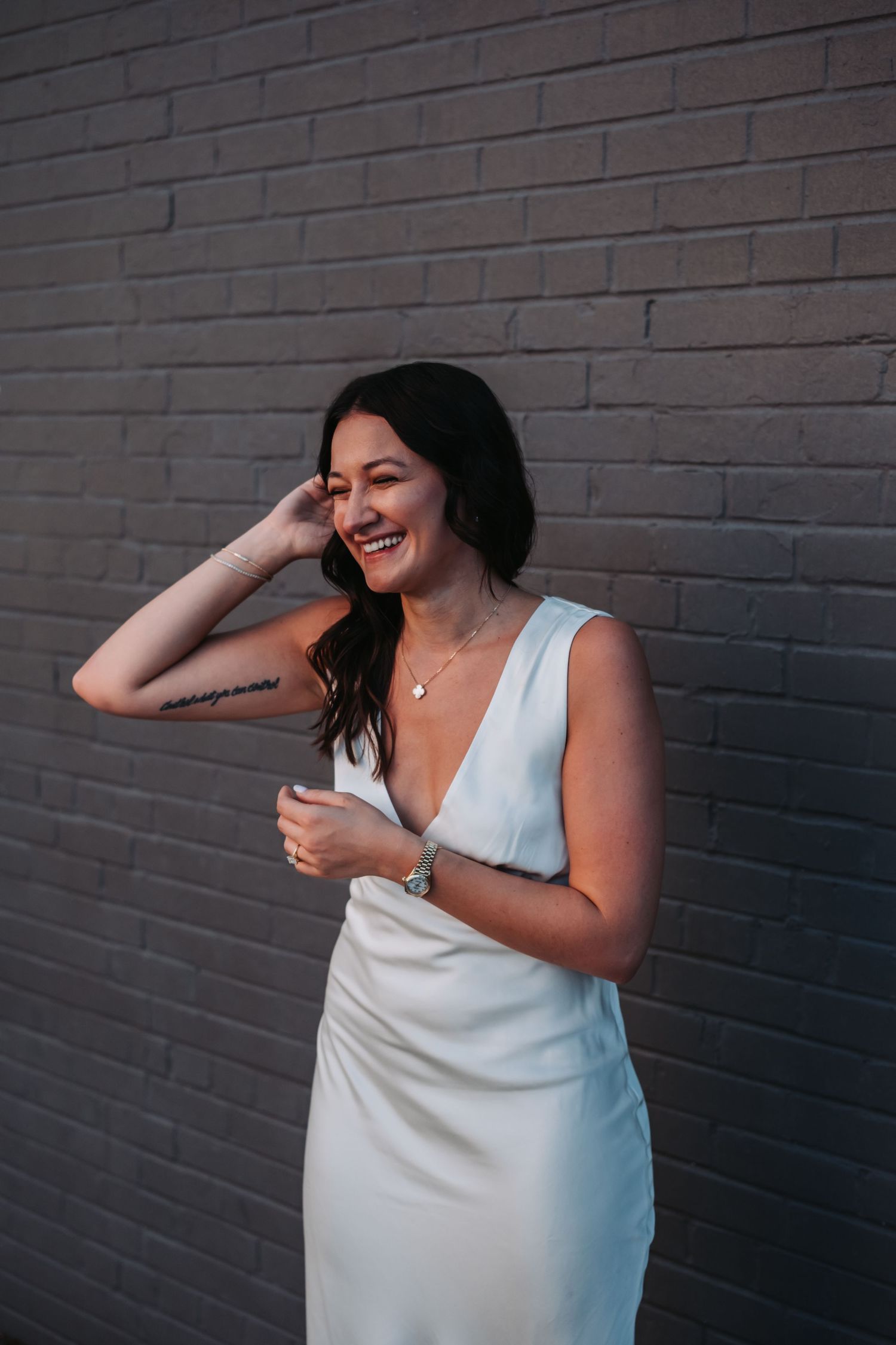 Laughing bride in white satin gown with tattoo posing against gray brick wall.
