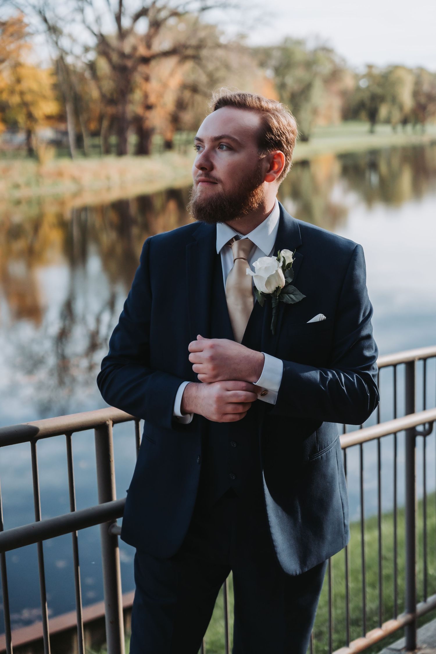 Dapper bearded groom in navy suit with white rose boutonniere poses by riverside at golden hour.