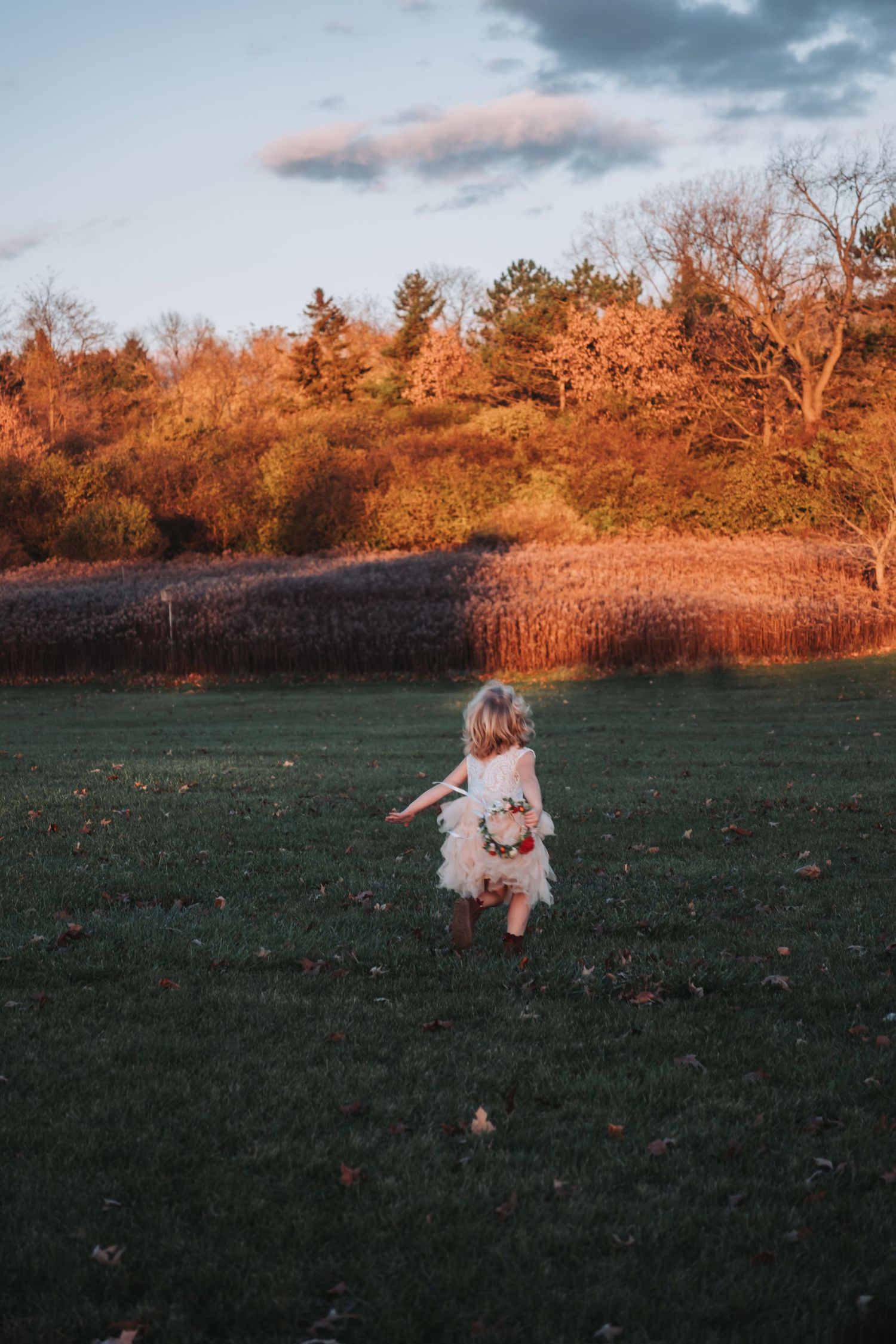 A young girl in a white dress runs across a green field holding flowers at golden hour sunset.