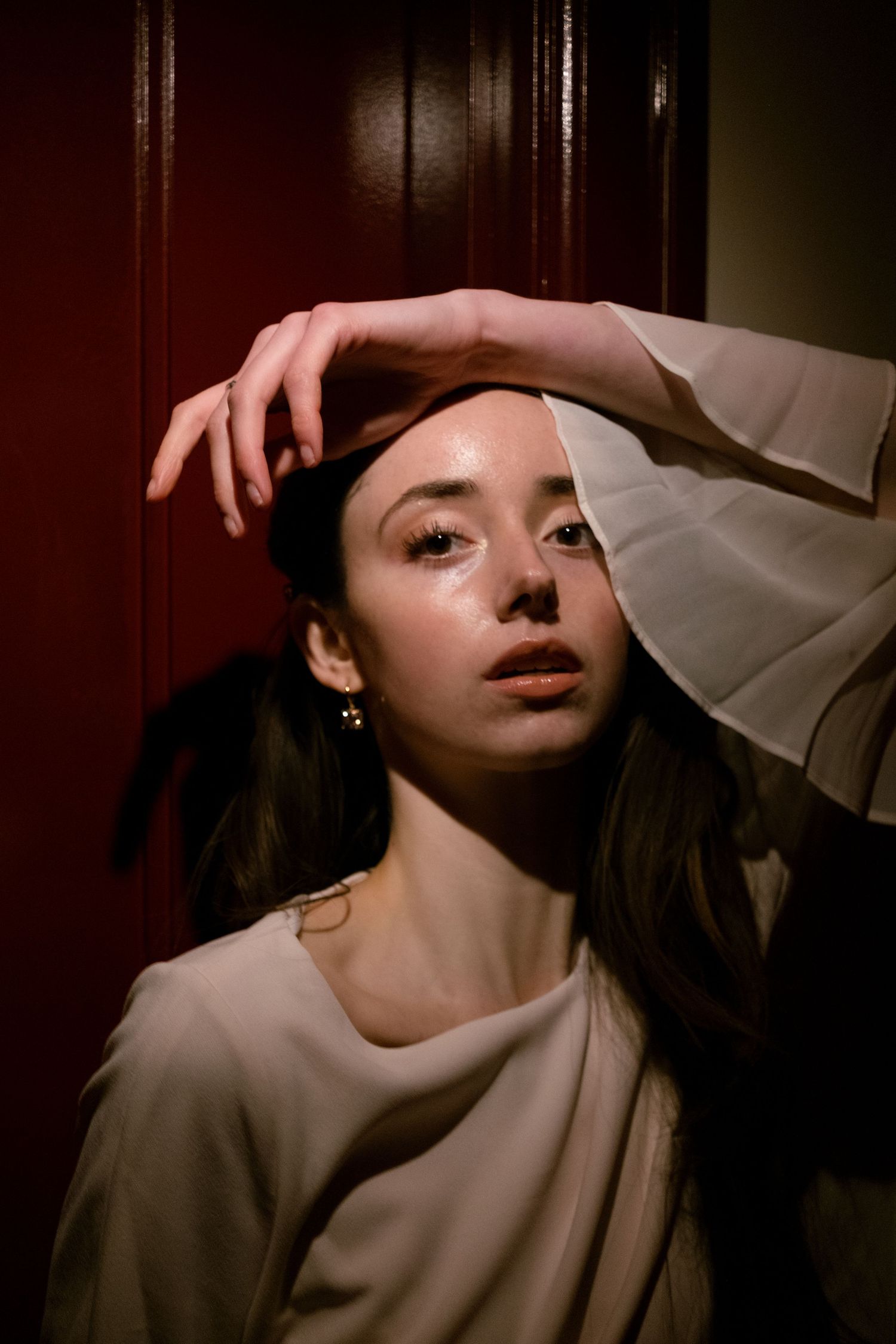 Young woman in white blouse poses dramatically against dark red door with hand raised above her head.