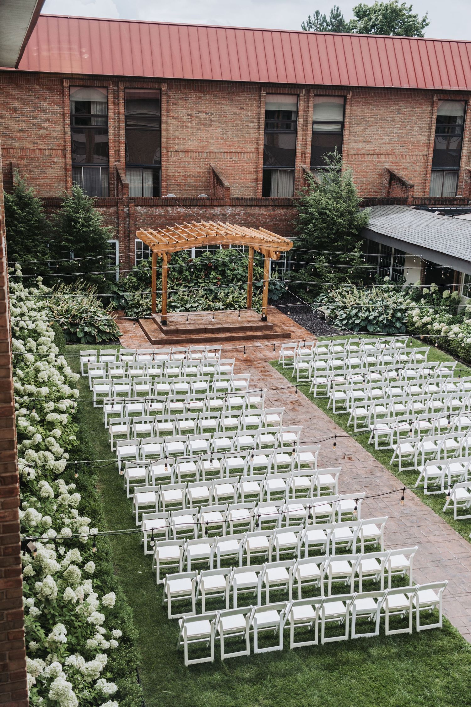 Outdoor wedding ceremony setup with white chairs and wooden pergola against brick building.
