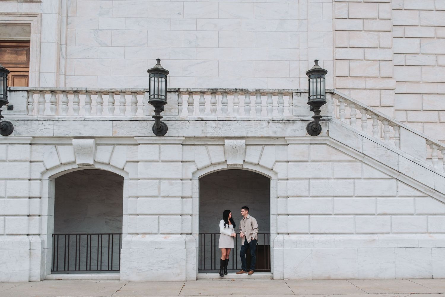 Couple standing together in an arched doorway of a grand white stone building with ornate lamp posts.