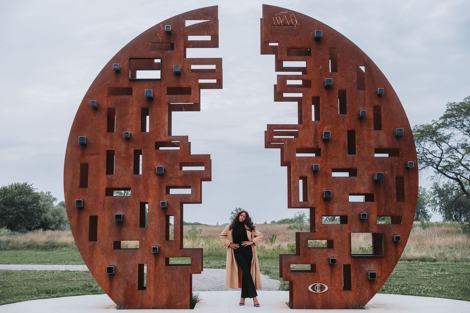 Woman posing between two large rusty metal circular sculptures with geometric cutouts outdoors.