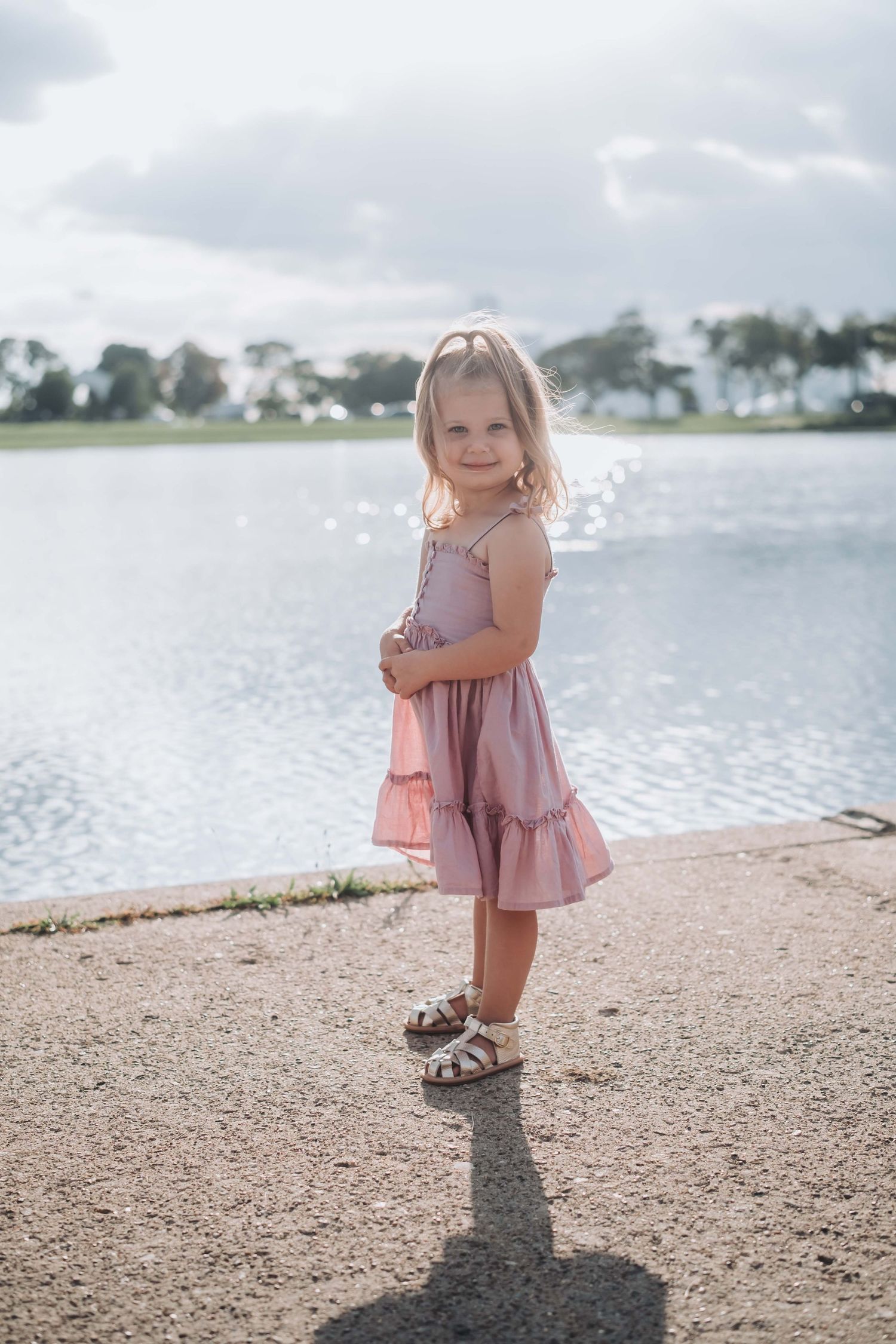 Young girl in pink dress and sandals holding a purse, standing by a lake on a sunny day.