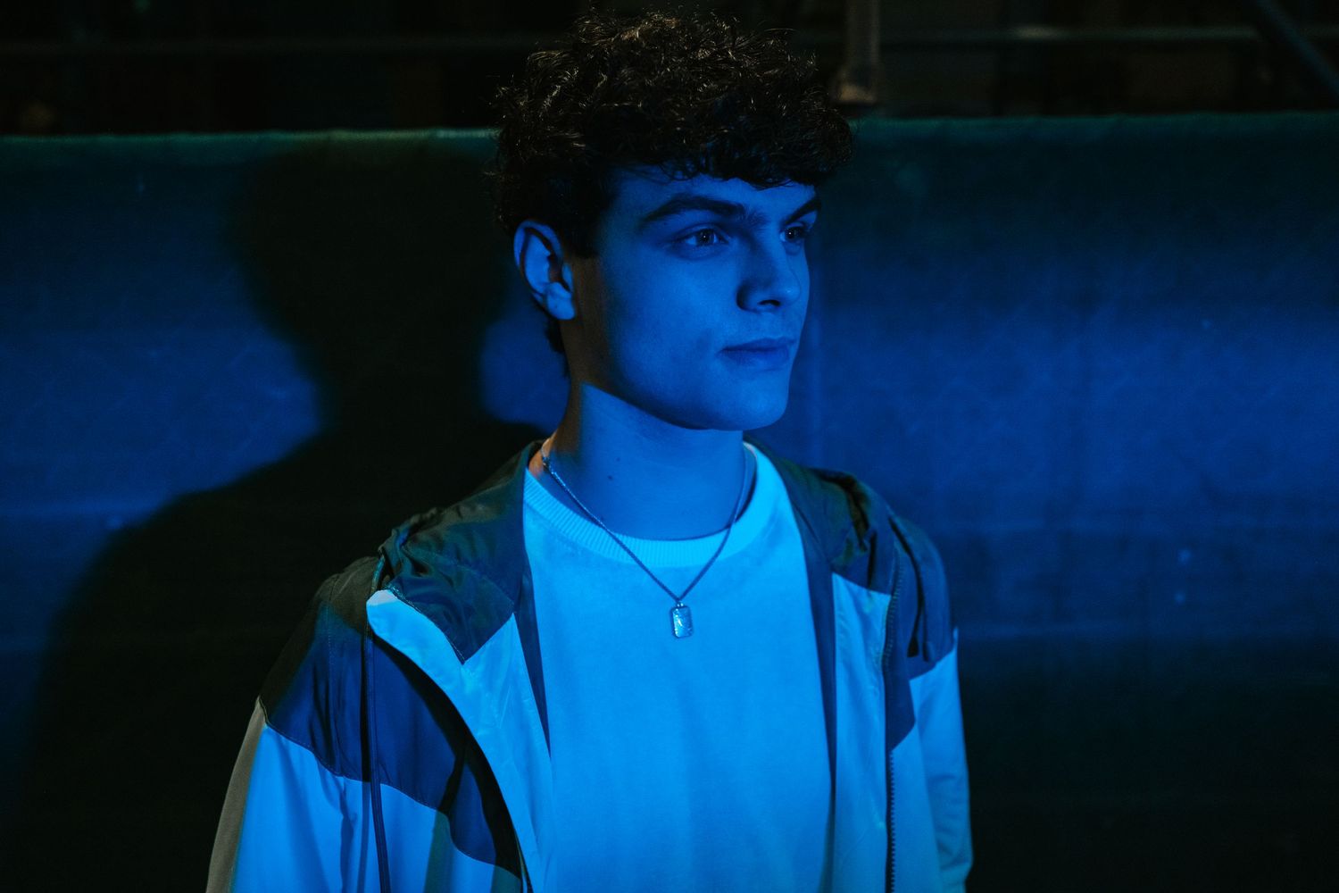Young man wearing a white shirt and jacket with a necklace, bathed in blue light against a dark background.