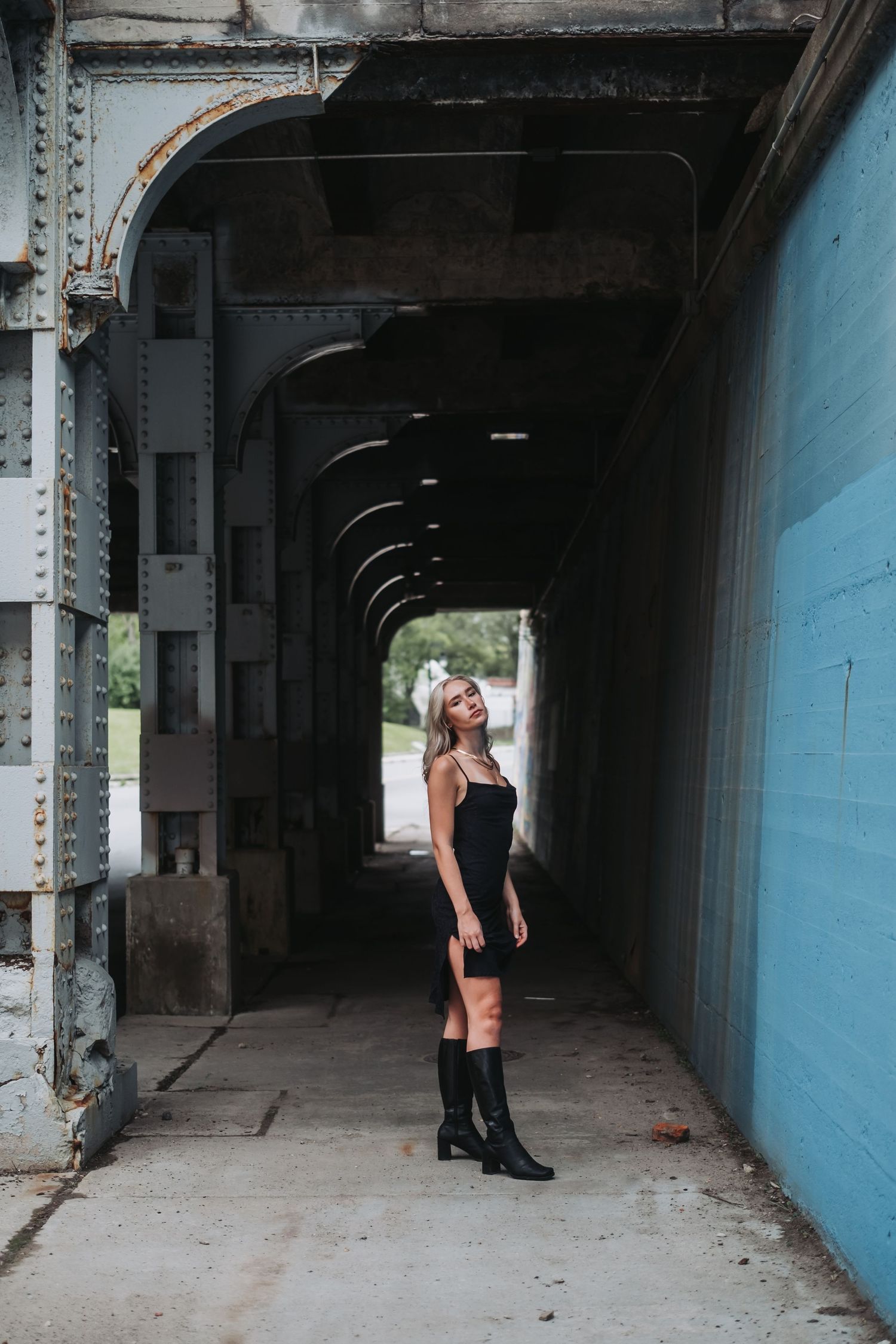 Woman in black mini dress and boots posing confidently under an urban bridge tunnel with arched architecture.