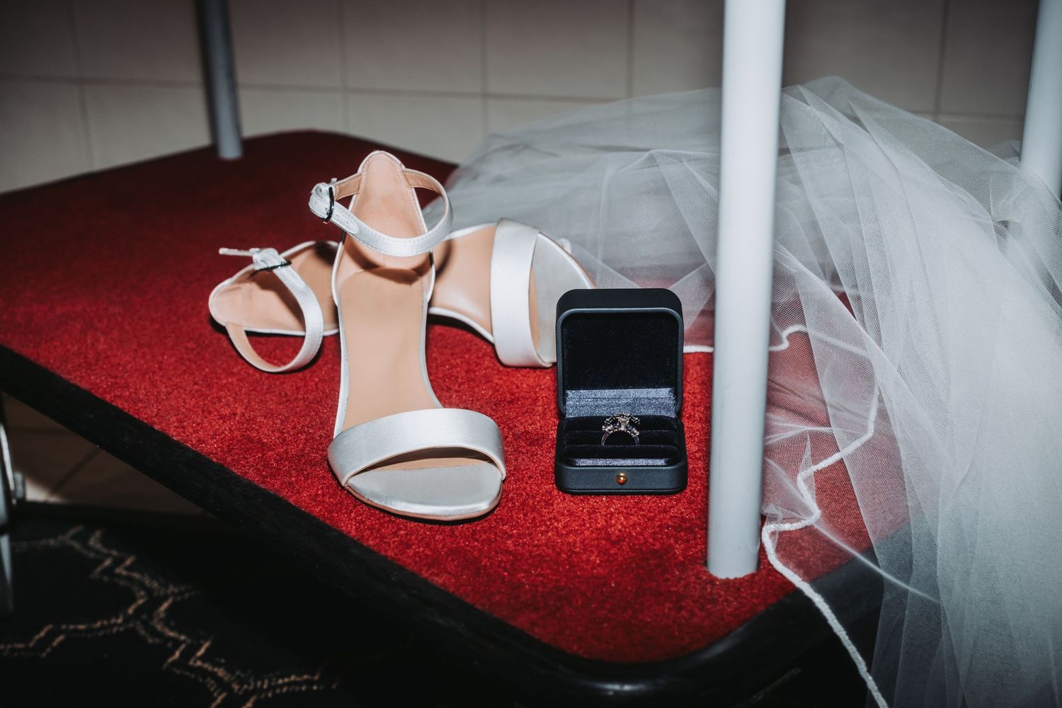 White bridal shoes and wedding ring box arranged on red carpet.