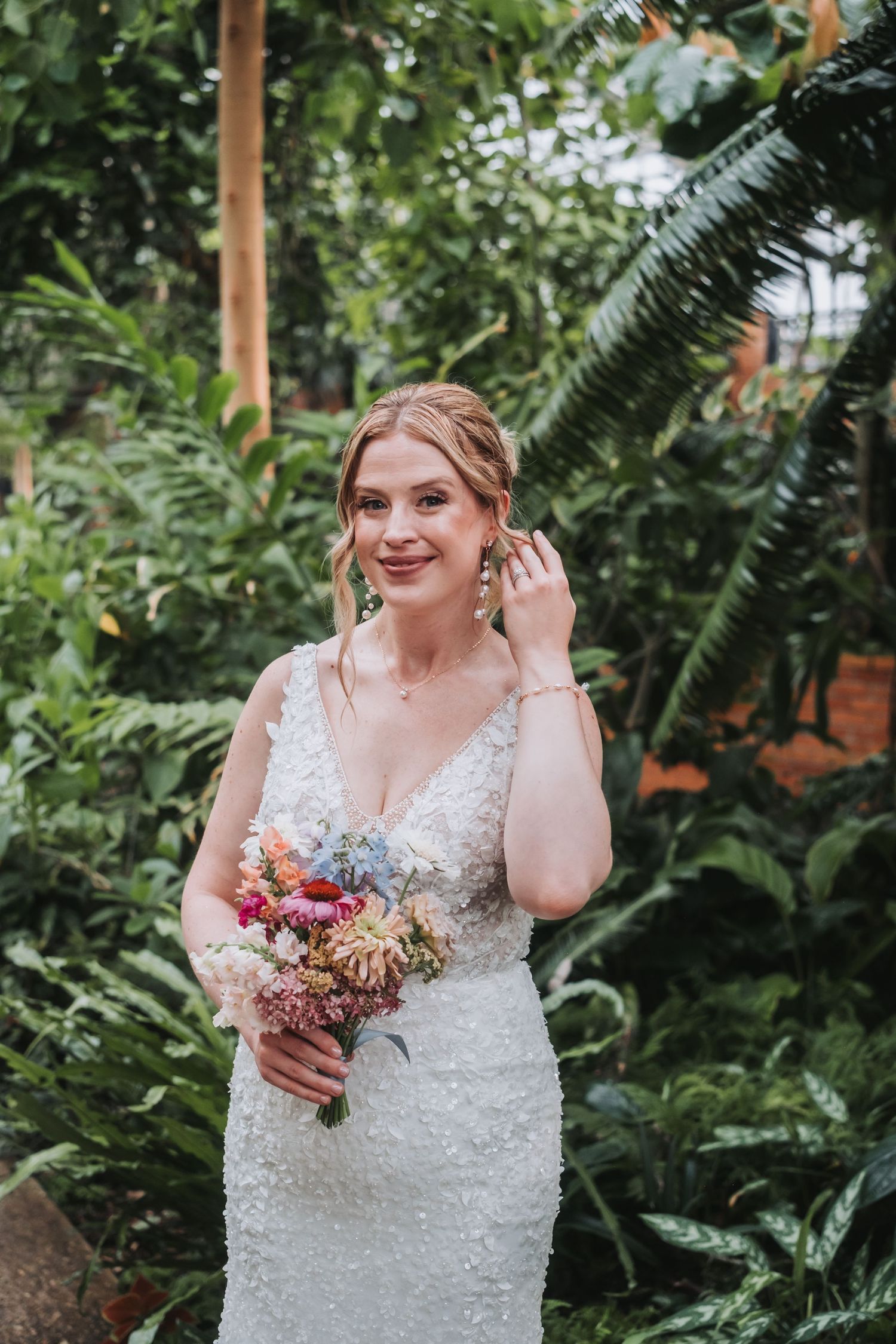 Person in white lace dress holding bouquet in lush greenhouse setting.