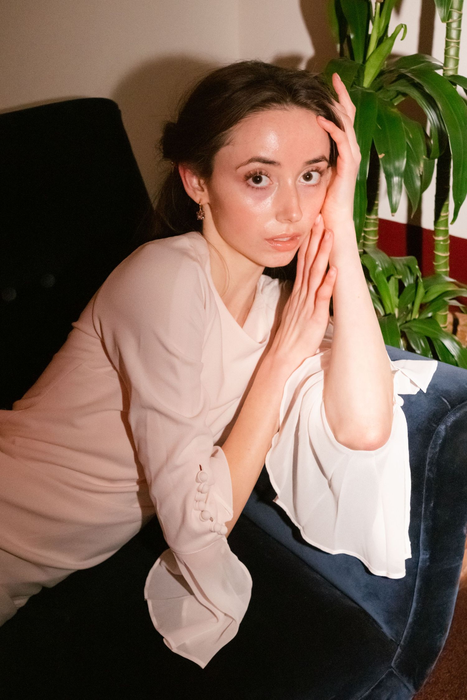 Young woman in pink blouse sitting on blue velvet chair, resting face in hands near indoor plant.