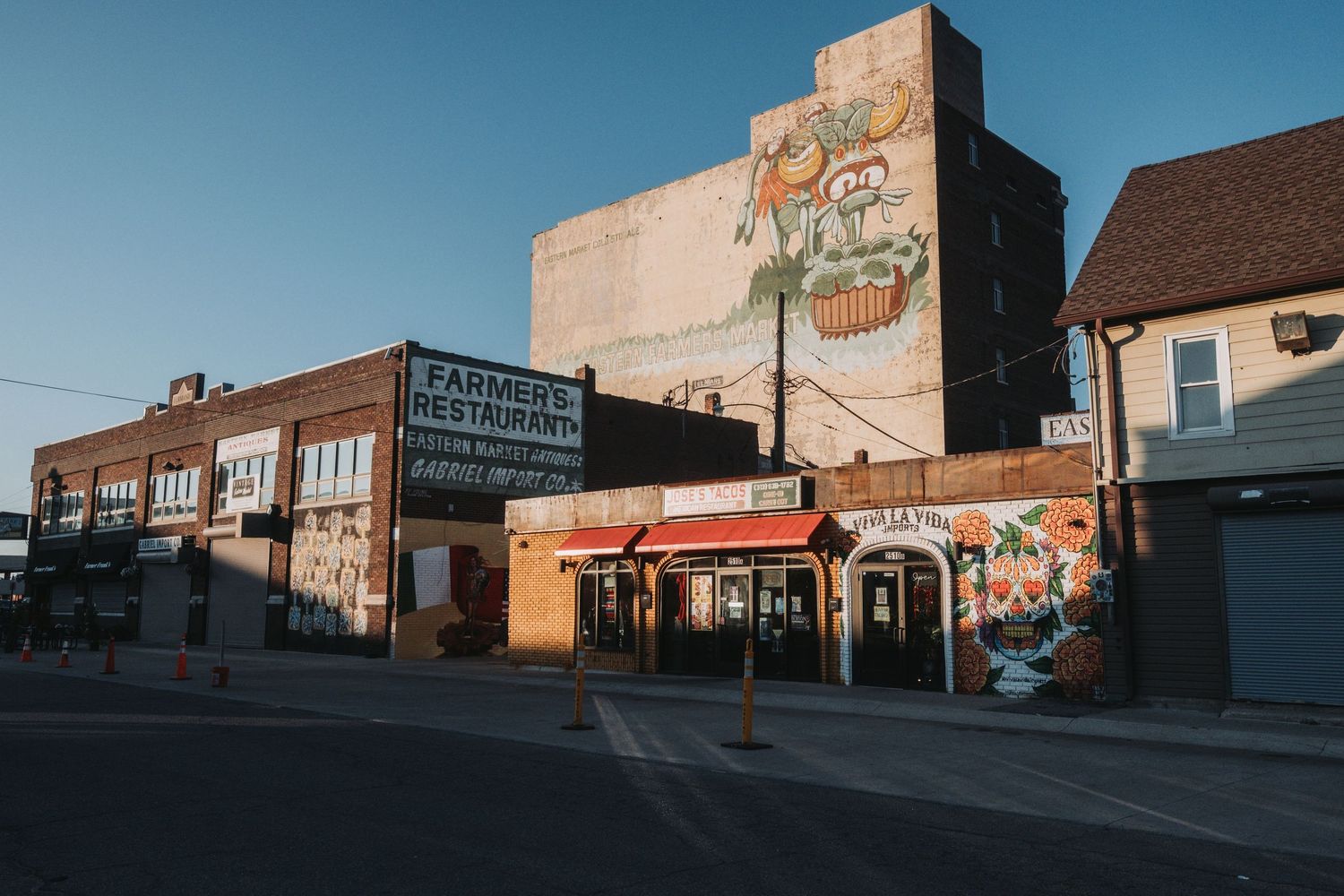 Urban street scene with colorful murals on brick buildings, including a farmers market sign, under a clear blue sky.